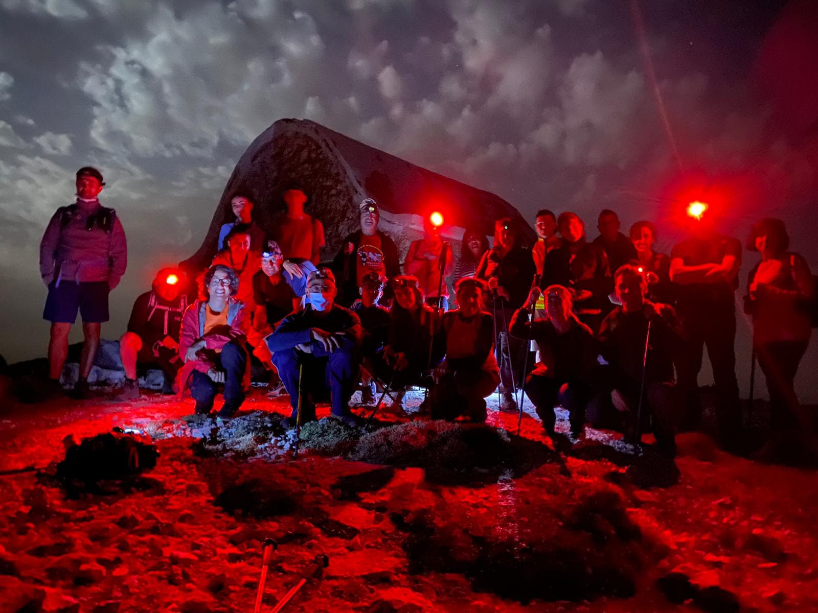 Un grupo se reúne en un entorno de montaña durante la noche, iluminado con luces frontales rojas bajo un cielo parcialmente cubierto.