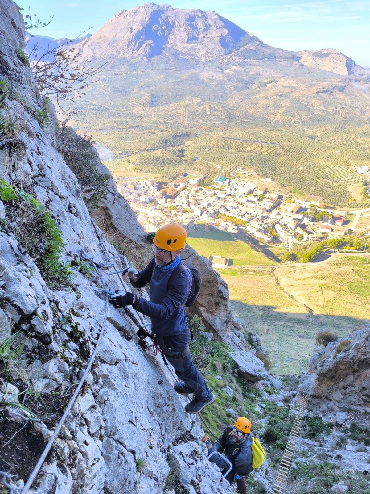 Un participante cruza en tirolina entre formaciones rocosas, suspendido sobre un paisaje de bosque mediterráneo y montañas.