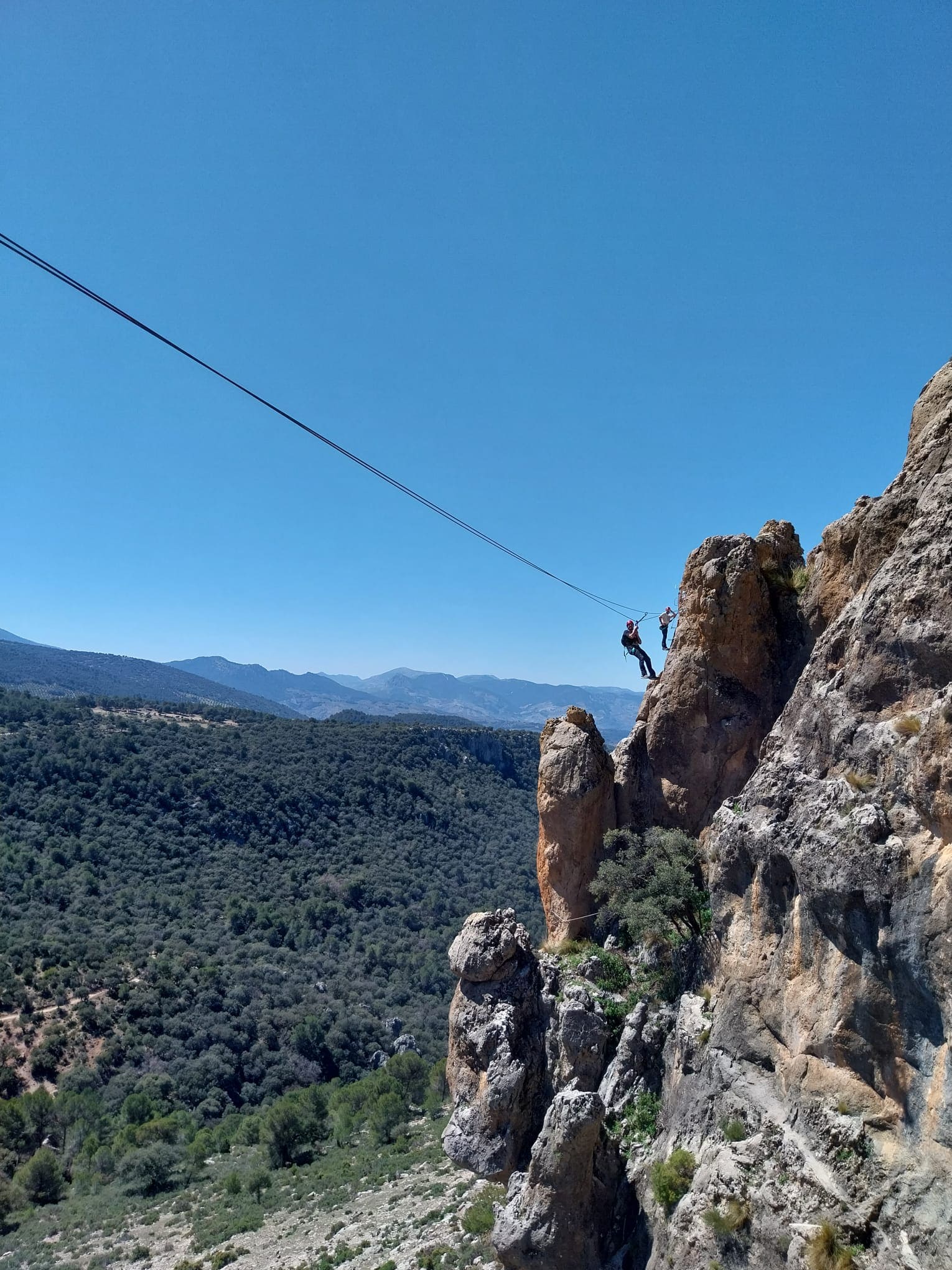 Una persona equipada con casco y arnés progresa por una pared rocosa, con amplias vistas a un pueblo y al paisaje de olivares característico de la zona.