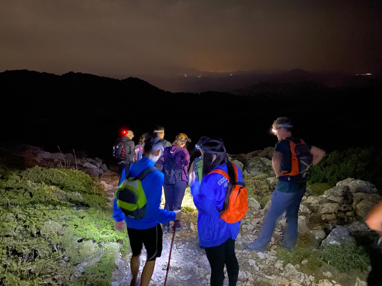 Varias personas caminan de noche por un sendero rocoso, iluminando el camino con linternas frontales mientras avanzan en grupo.