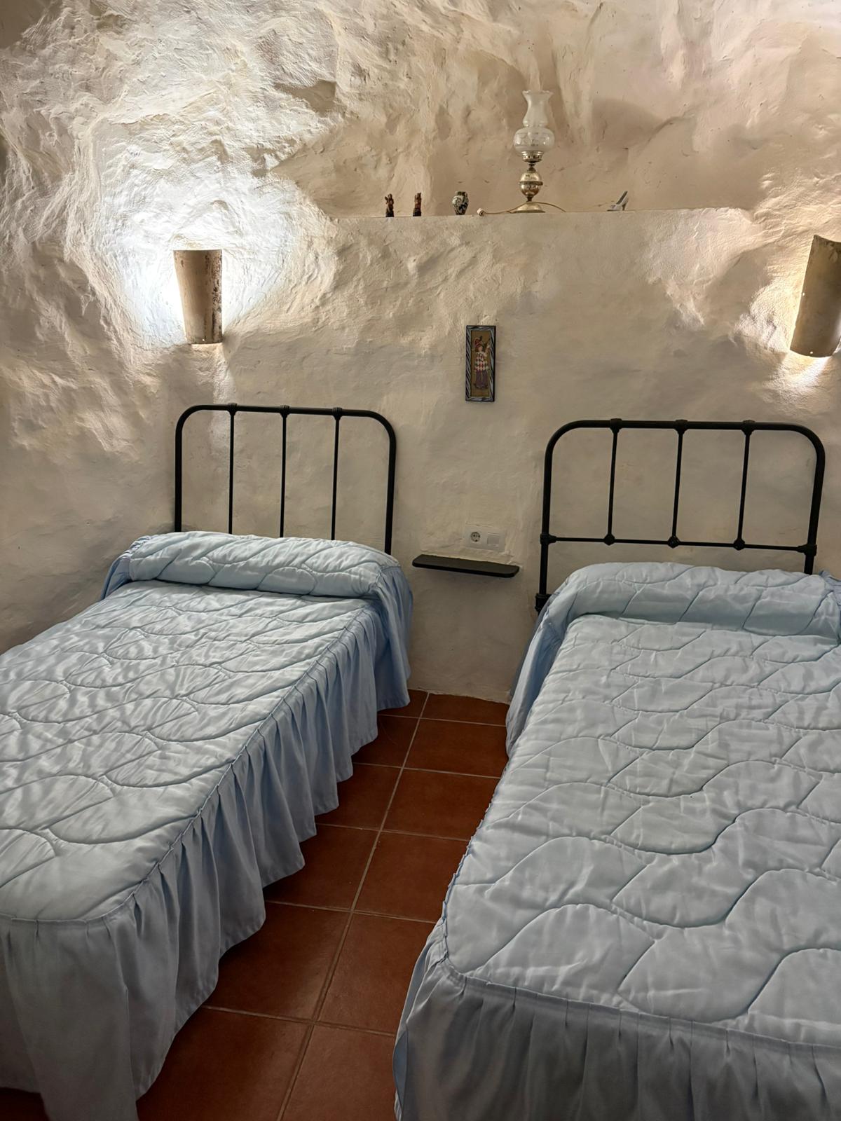 Habitación doble con camas individuales y cabeceros de forja, en un ambiente sencillo y funcional. La iluminación tenue resalta la textura natural de la roca.
