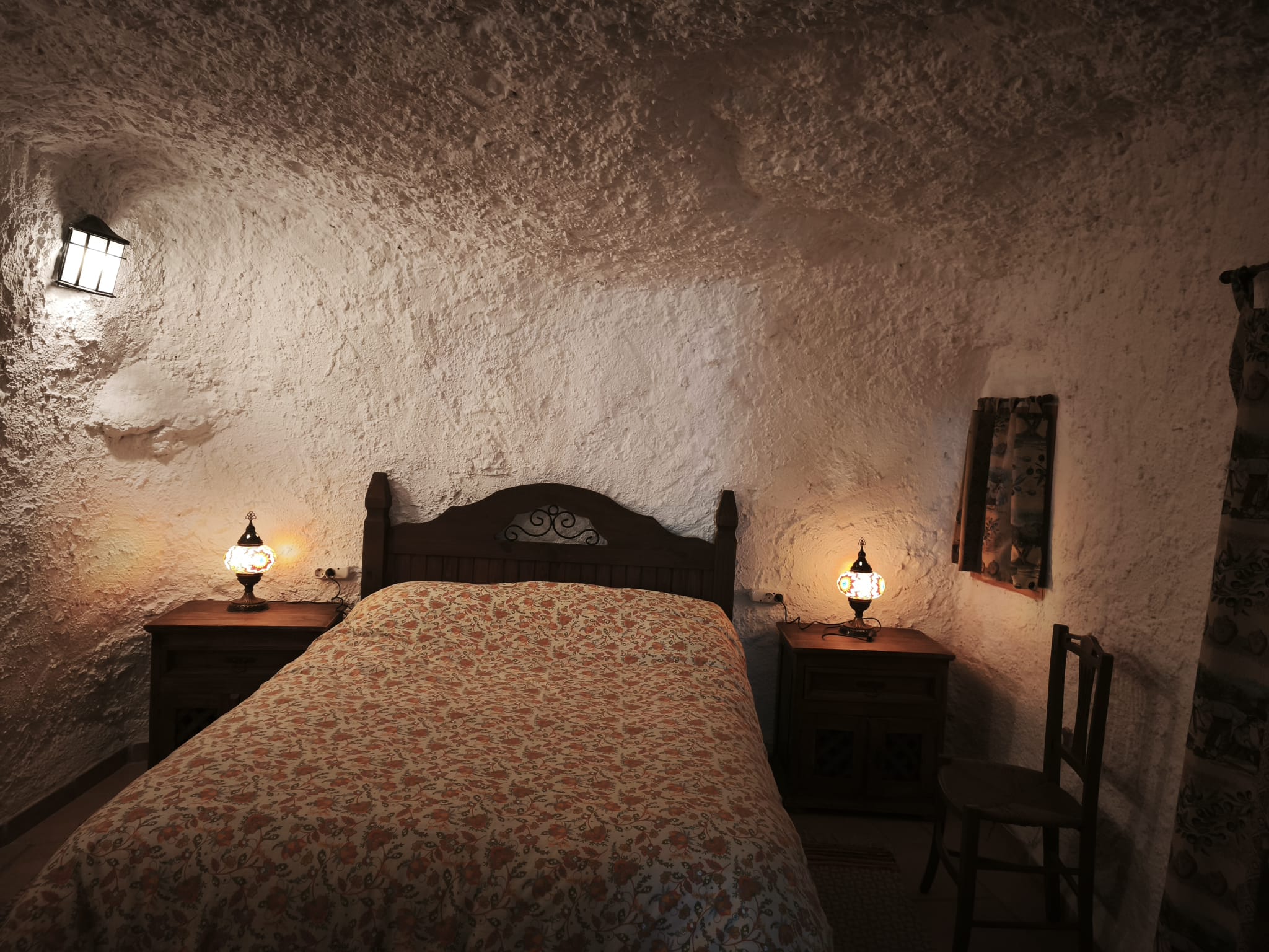 Dormitorio de ambiente íntimo, con cama de matrimonio y mesitas de madera. La luz tenue y las paredes de roca crean un entorno tranquilo ideal para el descanso.