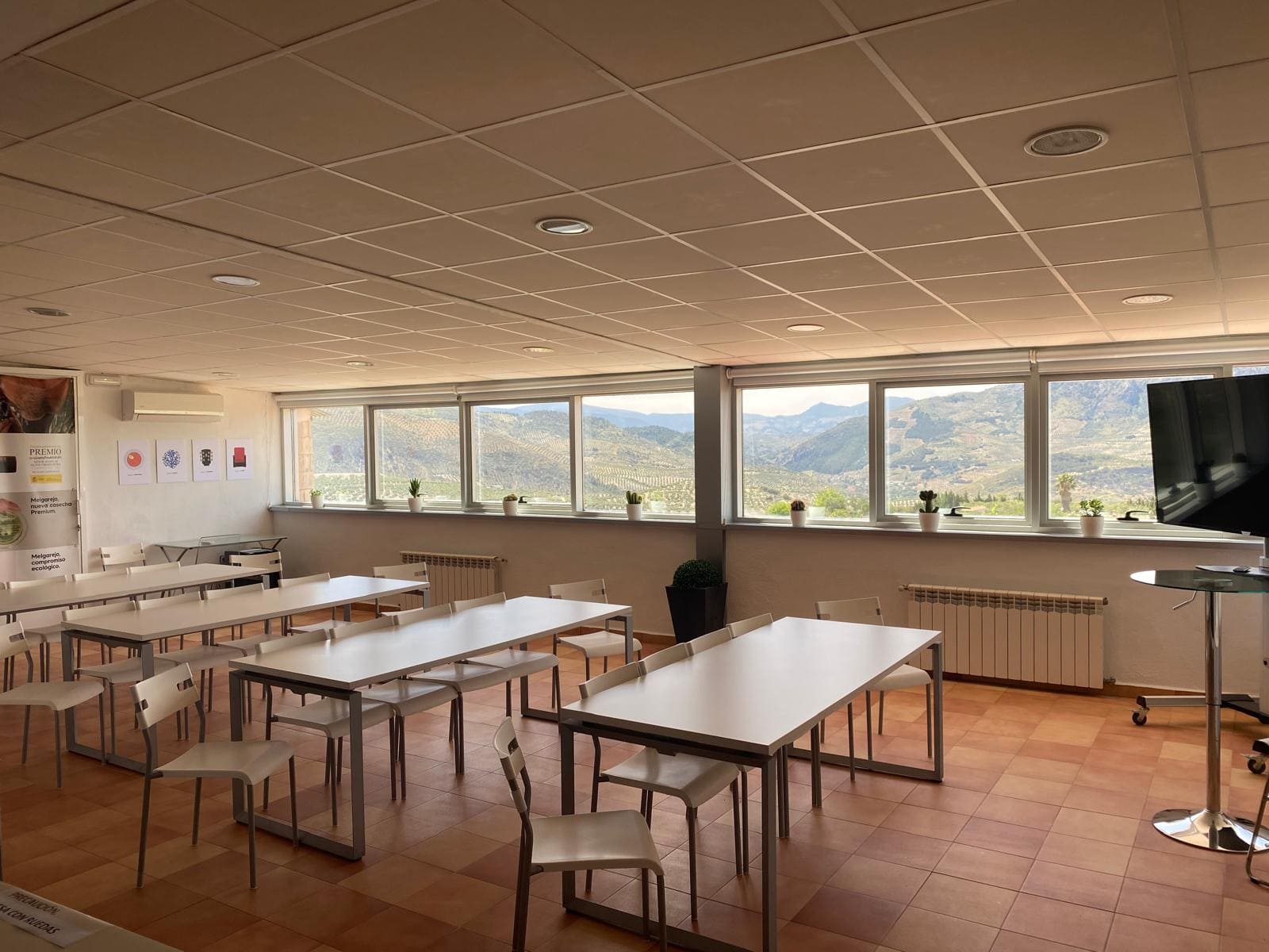 Sala interior con mesas y sillas preparadas para actividades formativas, con grandes ventanales y vistas a un paisaje montañoso.