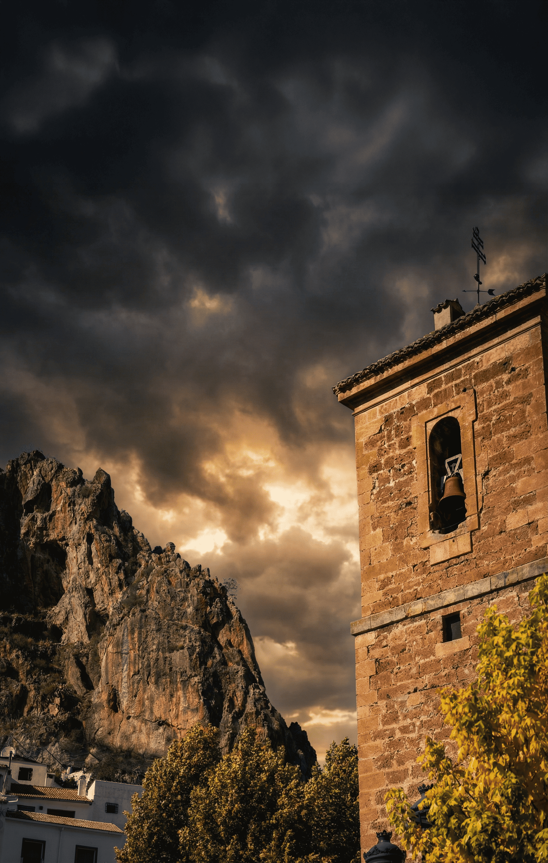 Campanario de piedra con una campana visible en su abertura, situado junto a una gran formación rocosa que se eleva sobre el paisaje. El cielo oscuro y cubierto de nubes dramáticas aporta una atmósfera intensa y de gran fuerza visual.