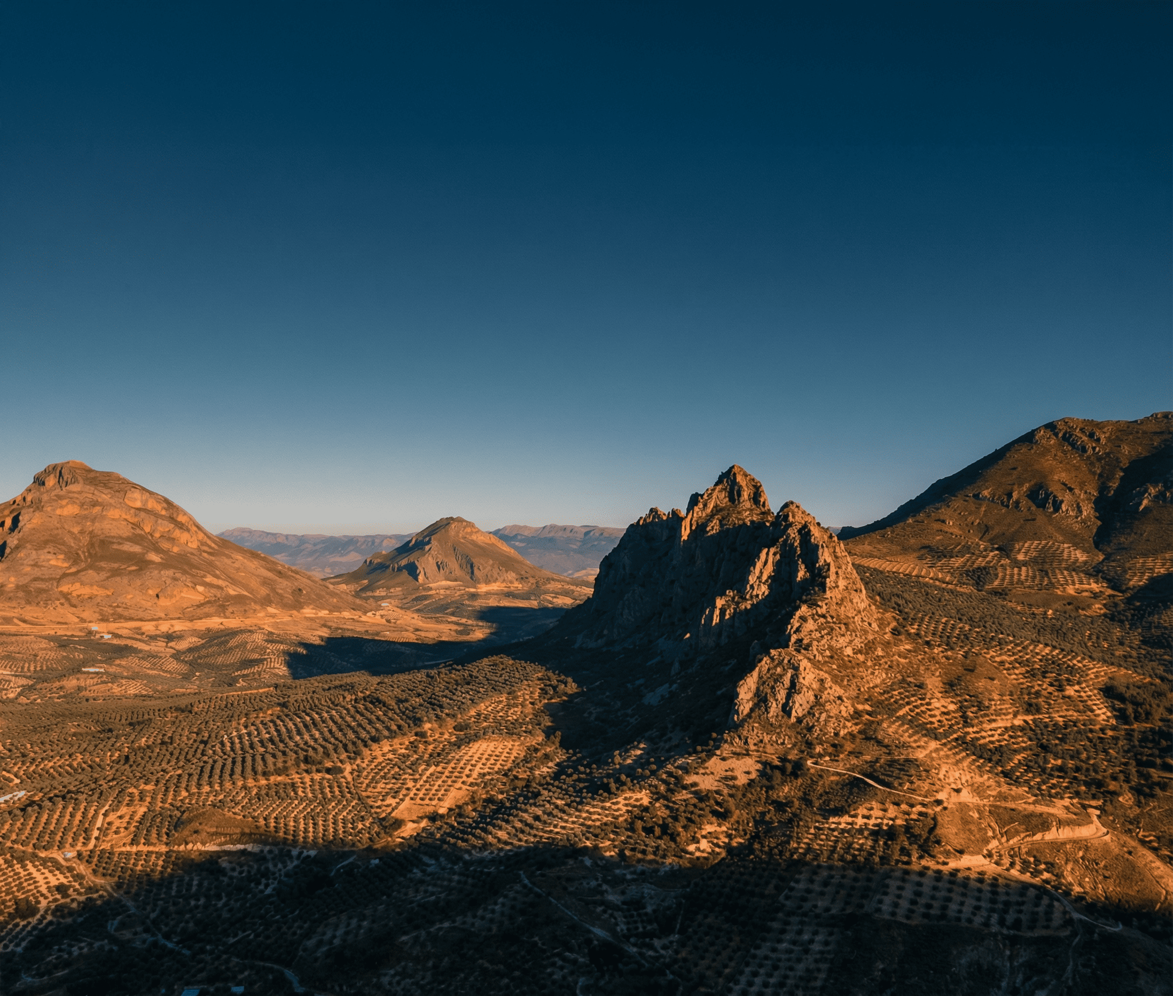 Paisaje montañoso de formas abruptas y tonos ocres, con una formación rocosa destacada en el centro rodeada de extensos olivares y laderas cultivadas. La luz baja proyecta sombras alargadas que realzan el relieve y la amplitud del entorno.
