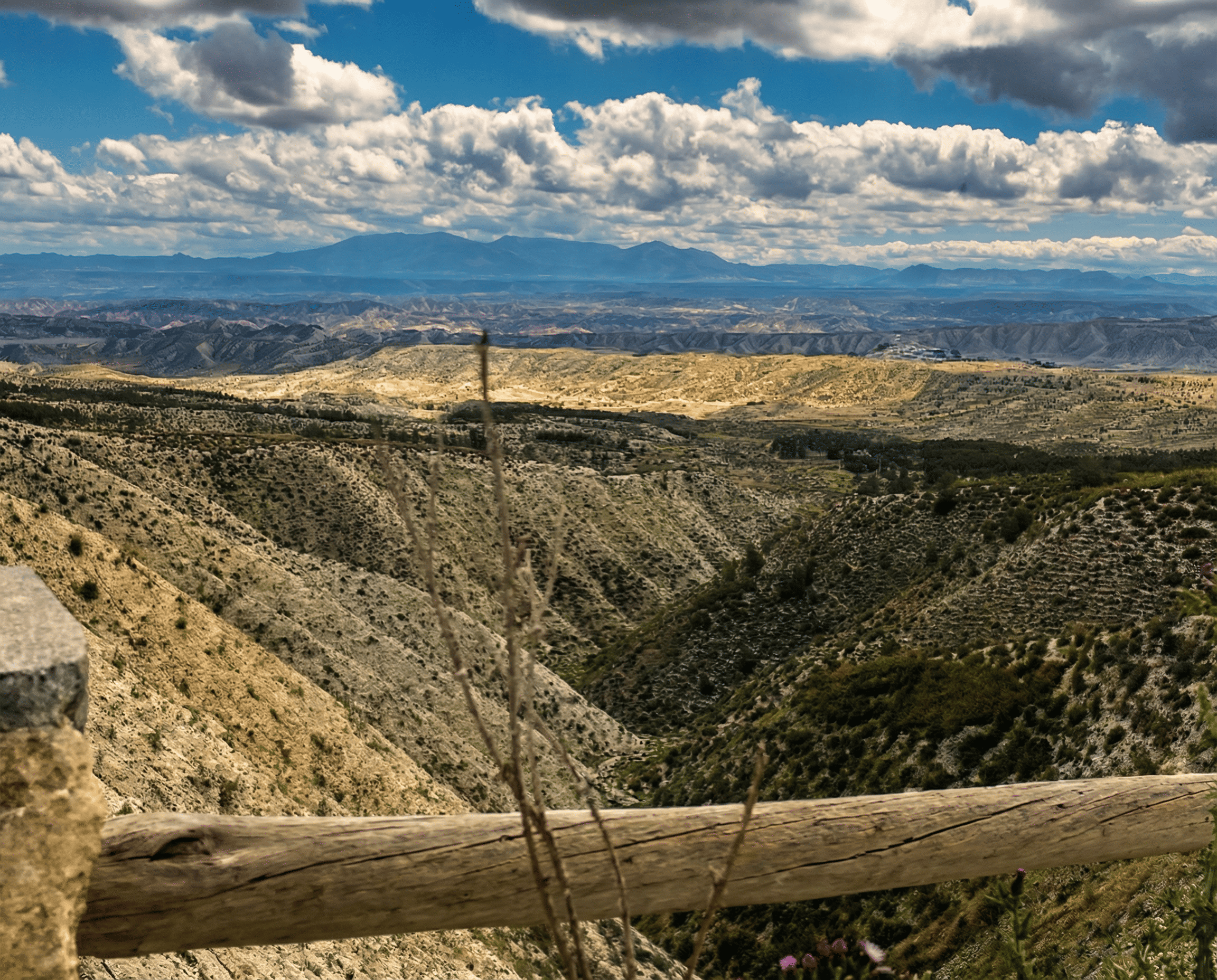 Vista panorámica de un paisaje de barrancos y lomas de tonos ocres, observado desde un mirador con barandilla de madera en primer plano. Al fondo se distinguen sierras lejanas bajo un cielo amplio cubierto de nubes densas y luminosas.