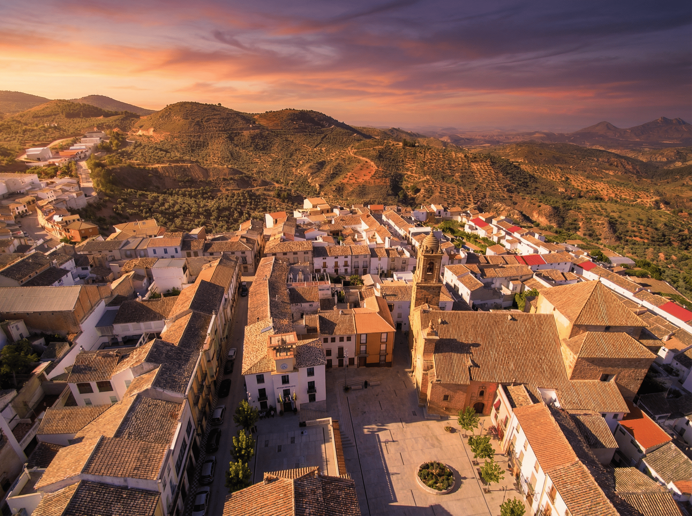 Vista aérea de un núcleo urbano de casas blancas y tejados de tonos tierra, organizado en torno a una plaza con iglesia y campanario. El pueblo se integra en un paisaje de colinas y olivares bajo un cielo de atardecer en tonos rosados y anaranjados.