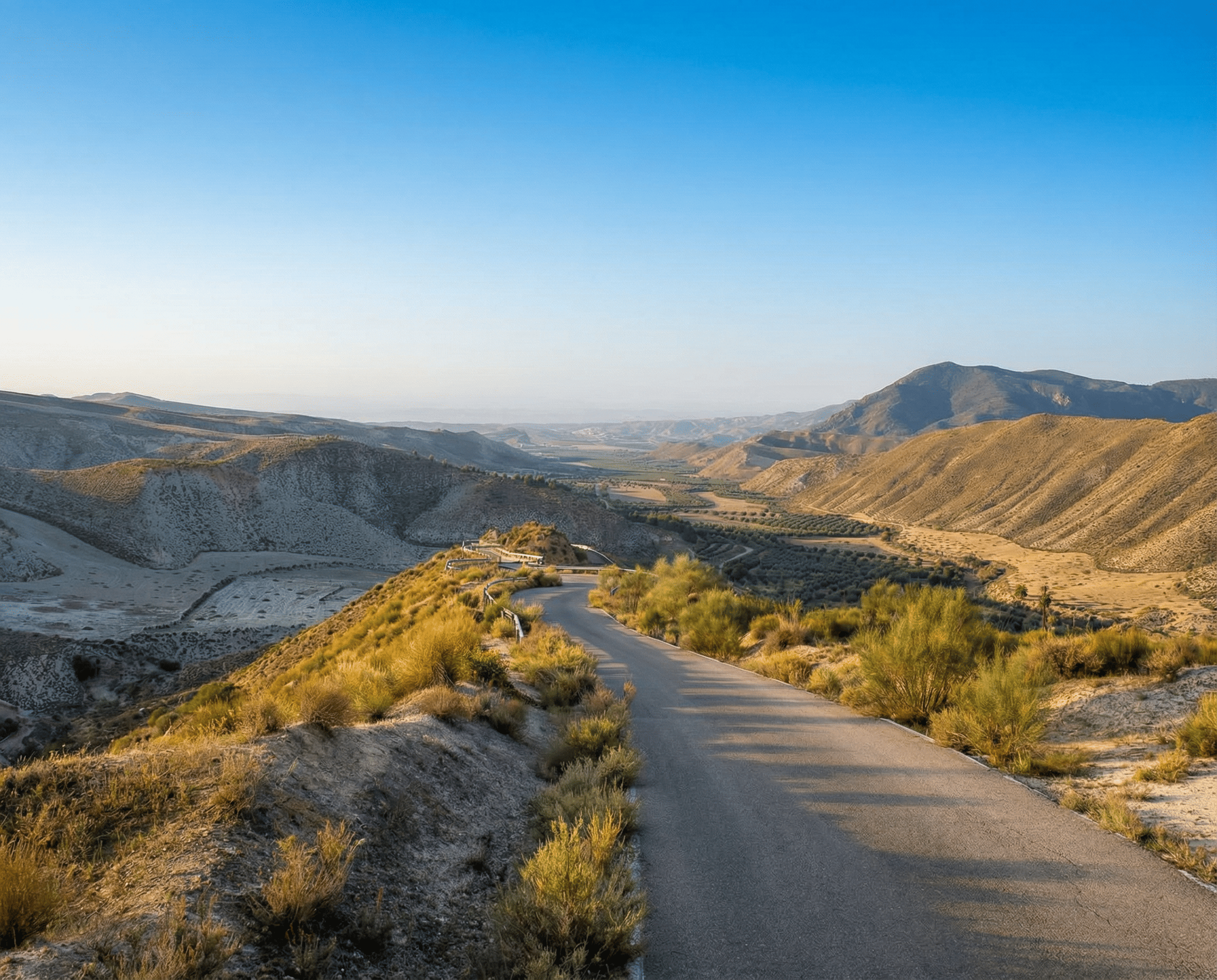 Carretera estrecha que desciende entre laderas de tonos ocres y vegetación dispersa, abriéndose hacia un amplio valle rodeado de montañas. La luz suave y el cielo despejado refuerzan la sensación de profundidad y amplitud del paisaje.