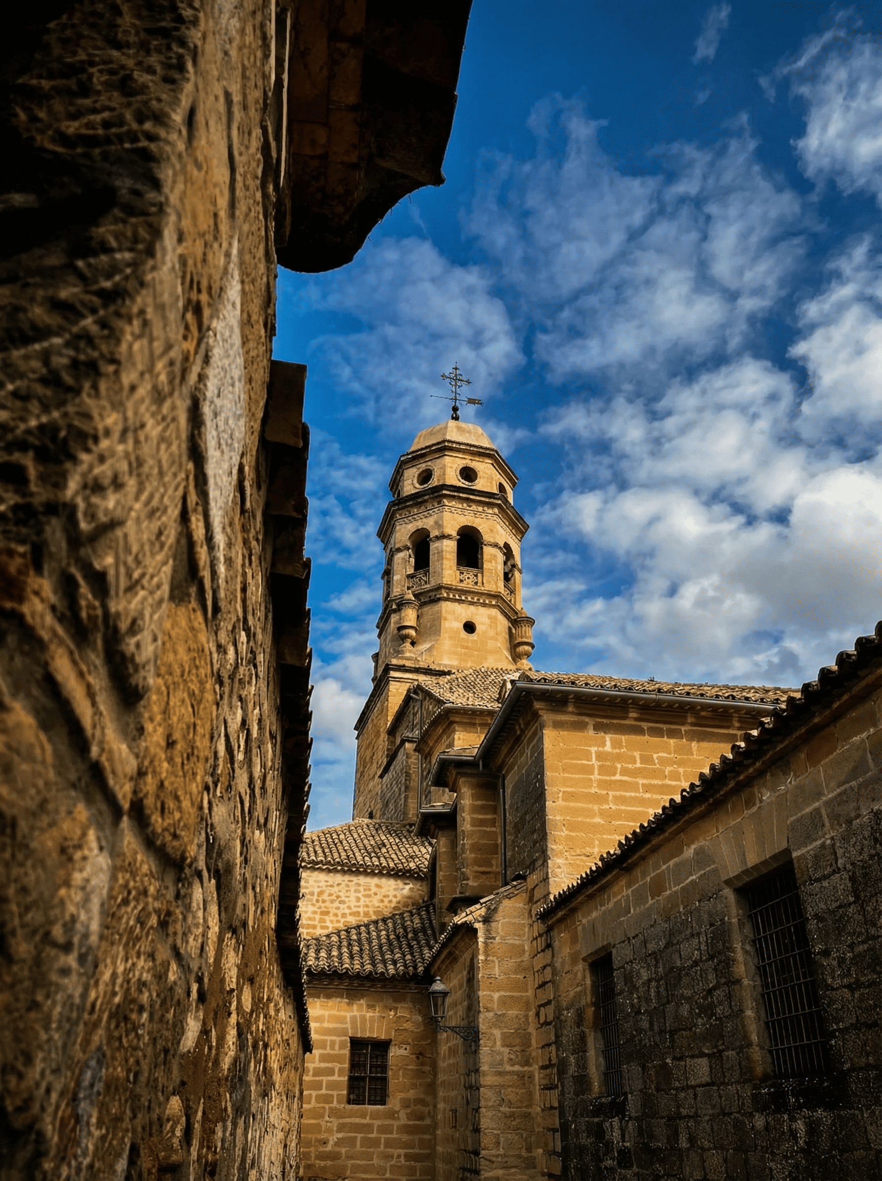 Un clocher en pierre vu depuis une ruelle étroite bordée de murs et de toits traditionnels. L'architecture aux teintes dorées se détache sur un ciel bleu parsemé de nuages blancs, conférant profondeur et verticalité à la scène.