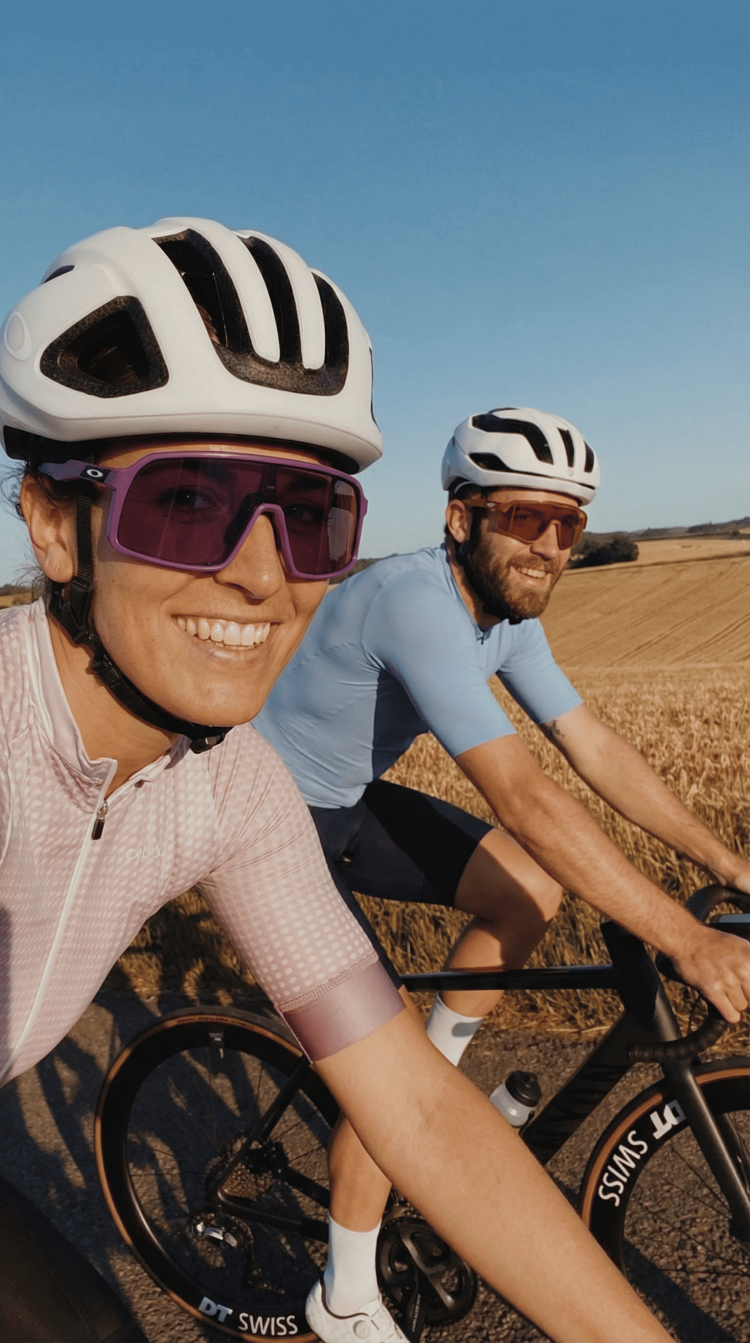 Deux cyclistes roulent sur une route bordée de champs à perte de vue ; tous deux portent un casque et des lunettes de cyclisme. Ce gros plan dégage une impression d'activité en plein air, de camaraderie et de plaisir à rouler.