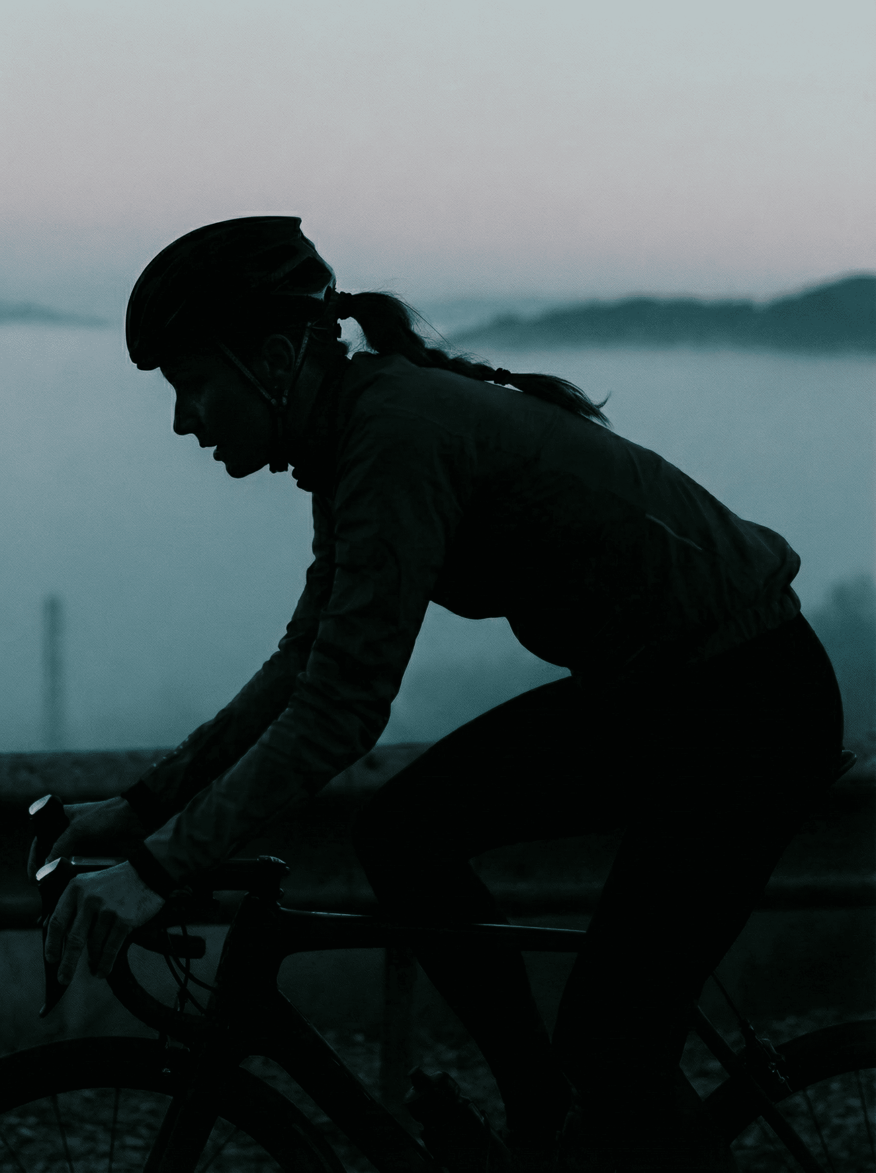 Perfil de una ciclista sobre bicicleta de carretera en un entorno de baja visibilidad, con niebla y luz tenue. La silueta oscura de la deportista destaca sobre un fondo difuminado, transmitiendo esfuerzo, concentración y ambiente de primera hora del día.