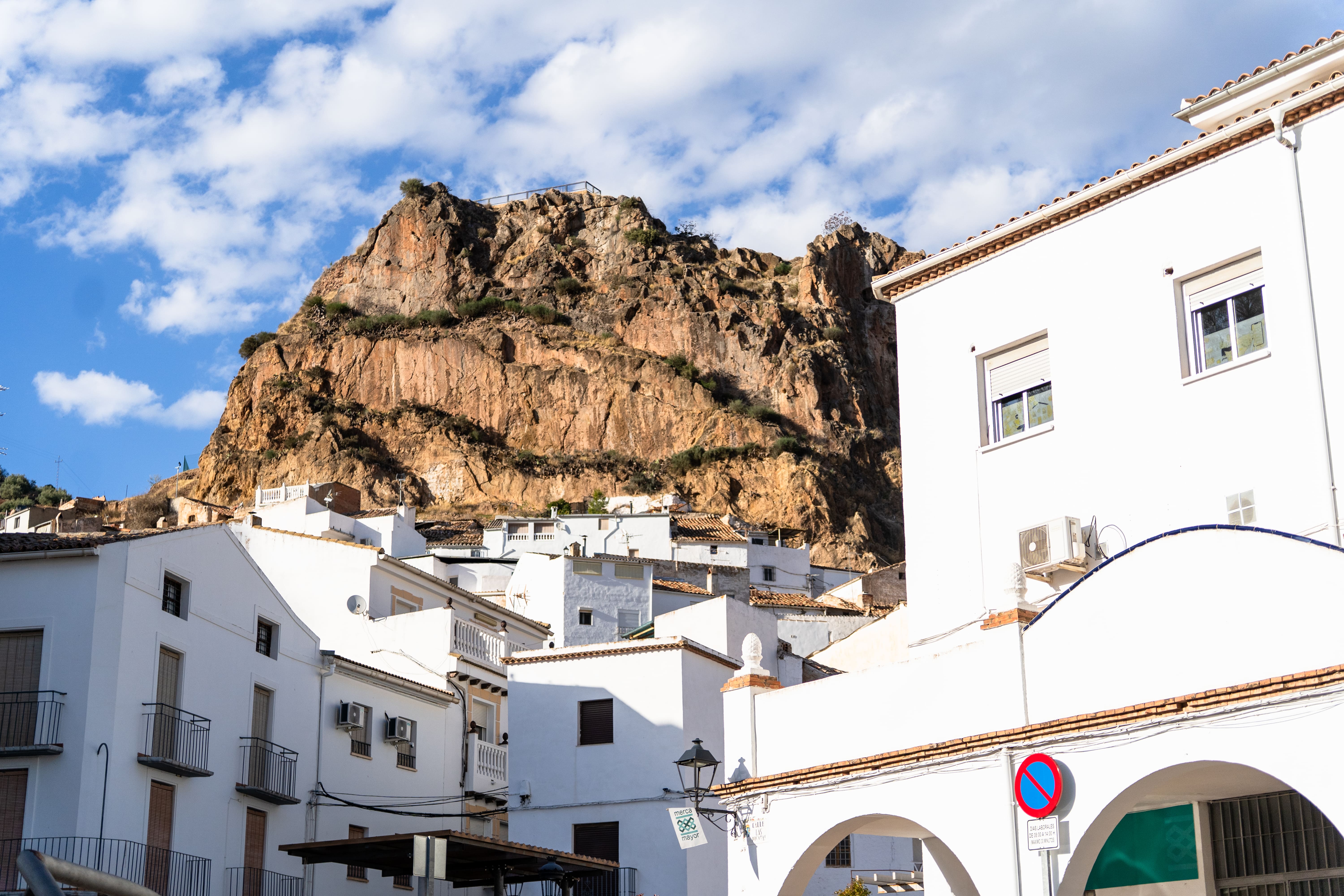 Primer plano del gran peñón rocoso que domina el pueblo, con paredes verticales de piedra en tonos anaranjados y marrones. En la parte inferior se aprecian casas blancas con tejados de teja, y a la derecha parte de una fachada blanca en primer plano. El cielo está despejado con nubes blancas.