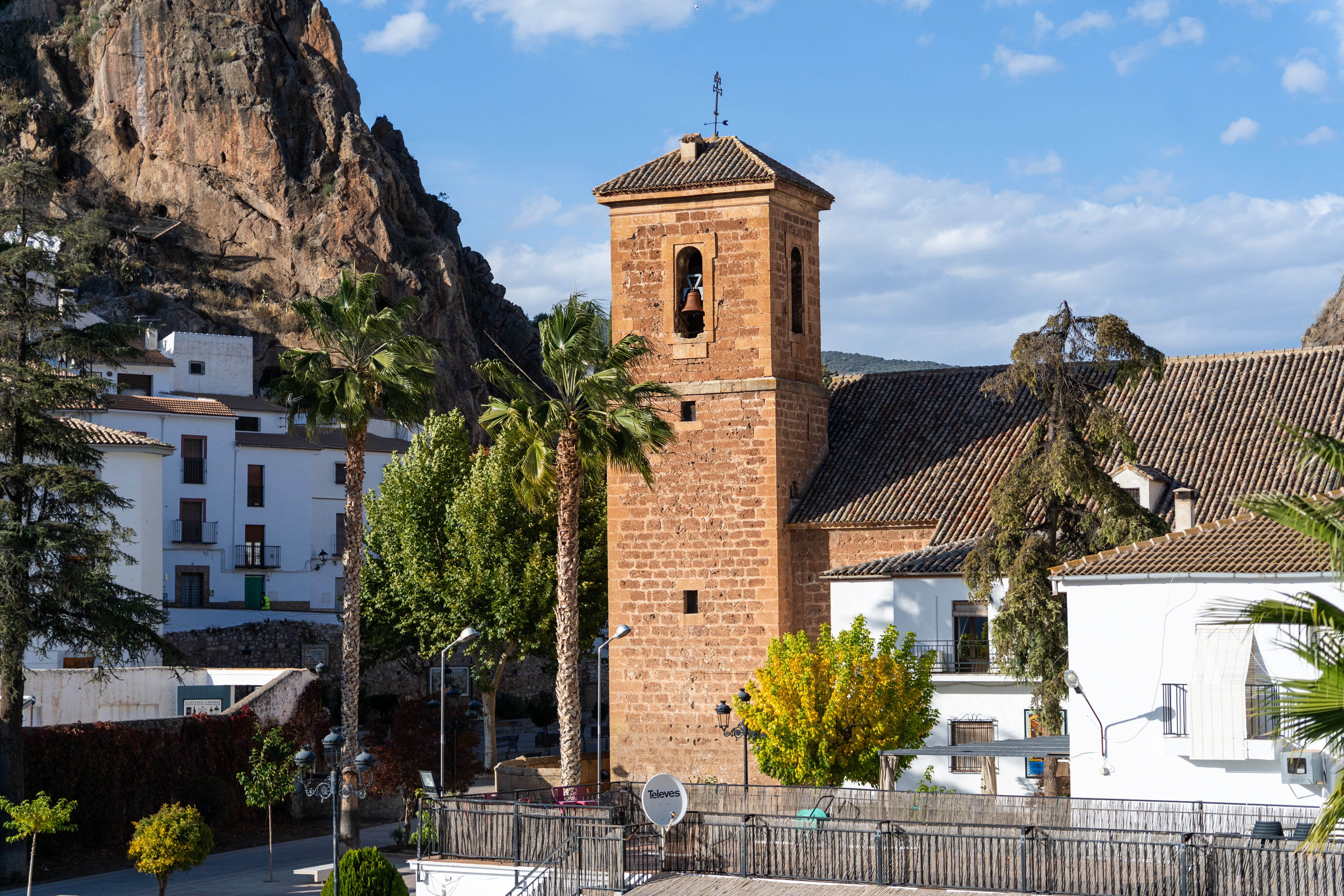 Vista general de Cambil en un día despejado: en primer plano aparece el campanario cuadrado de piedra rojiza de la Iglesia de Nuestra Señora de la Encarnación, con la campana visible. A su alrededor se ven palmeras, tejados de teja y algunas casas blancas del pueblo. Al fondo destaca una gran peña rocosa que domina el paisaje.