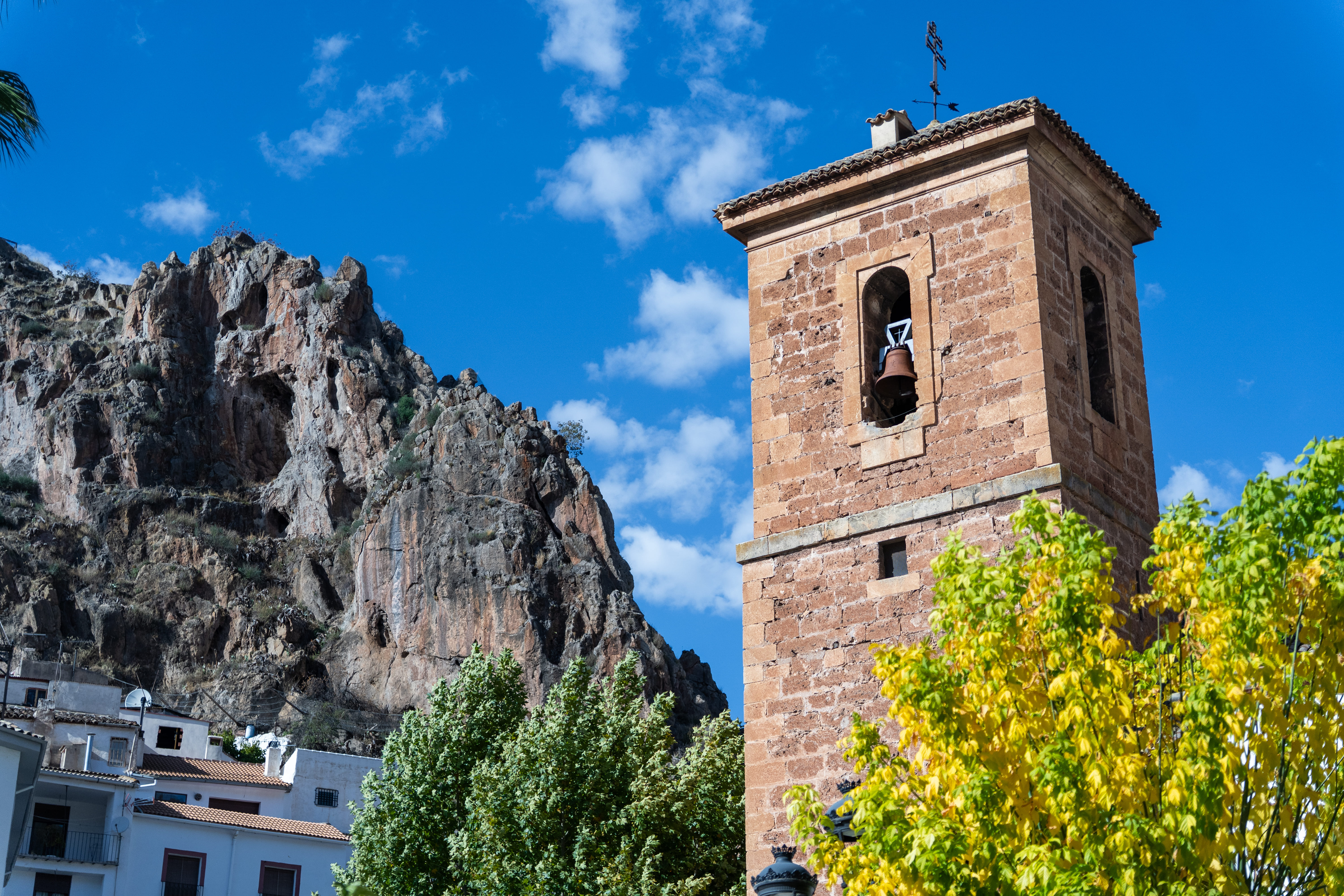 Detalle del campanario en primer plano, con la campana visible, y detrás una ladera rocosa muy escarpada. En la parte inferior se insinúan tejados y fachadas blancas del pueblo, parcialmente ocultas por las copas de los árboles. El cielo aparece azul con algunas nubes pequeñas.