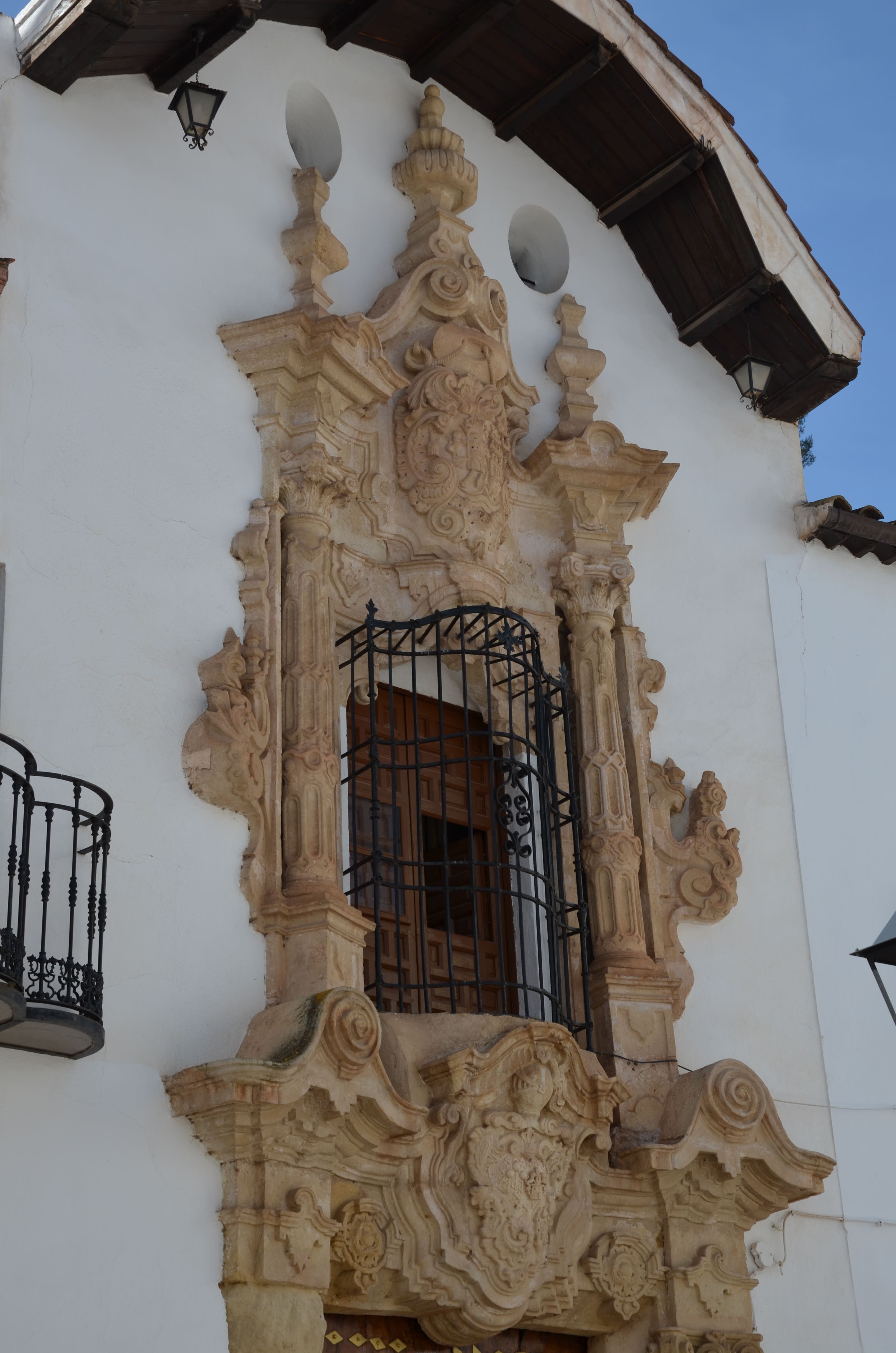 Fachada blanca de dos plantas con balcones de hierro forjado, puerta de madera y una hornacina con una pequeña figura religiosa en la esquina.