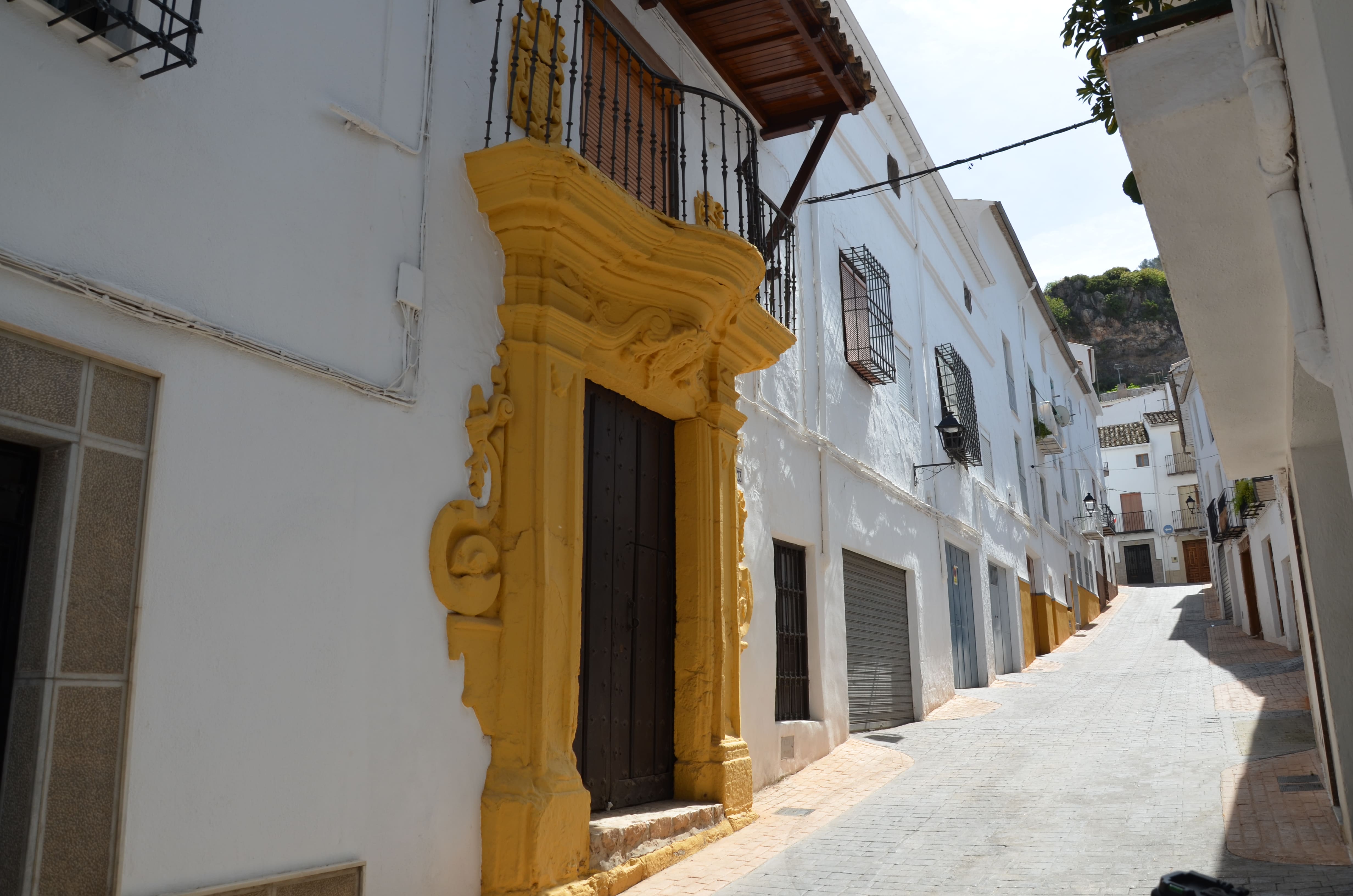 Calle estrecha con casas blancas; en primer plano destaca una portada barroca amarilla con balcón de forja encima, y al fondo se ve la calle en cuesta bajo un cielo azul.