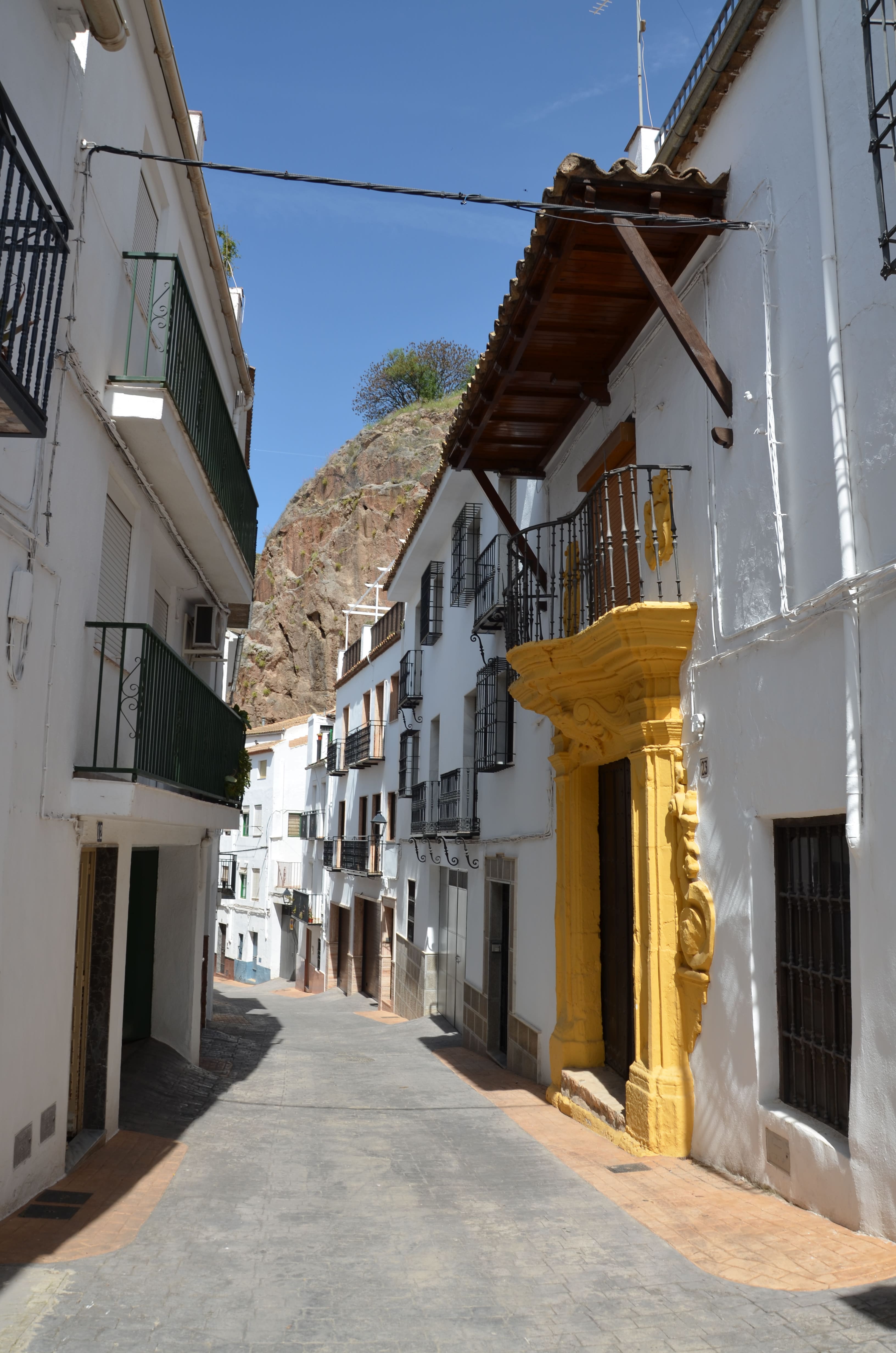 Calle angosta entre fachadas blancas con balcones y rejas; en primer plano aparece un edificio con gran alero de madera y detalles amarillos en la fachada, con una peña rocosa al fondo.