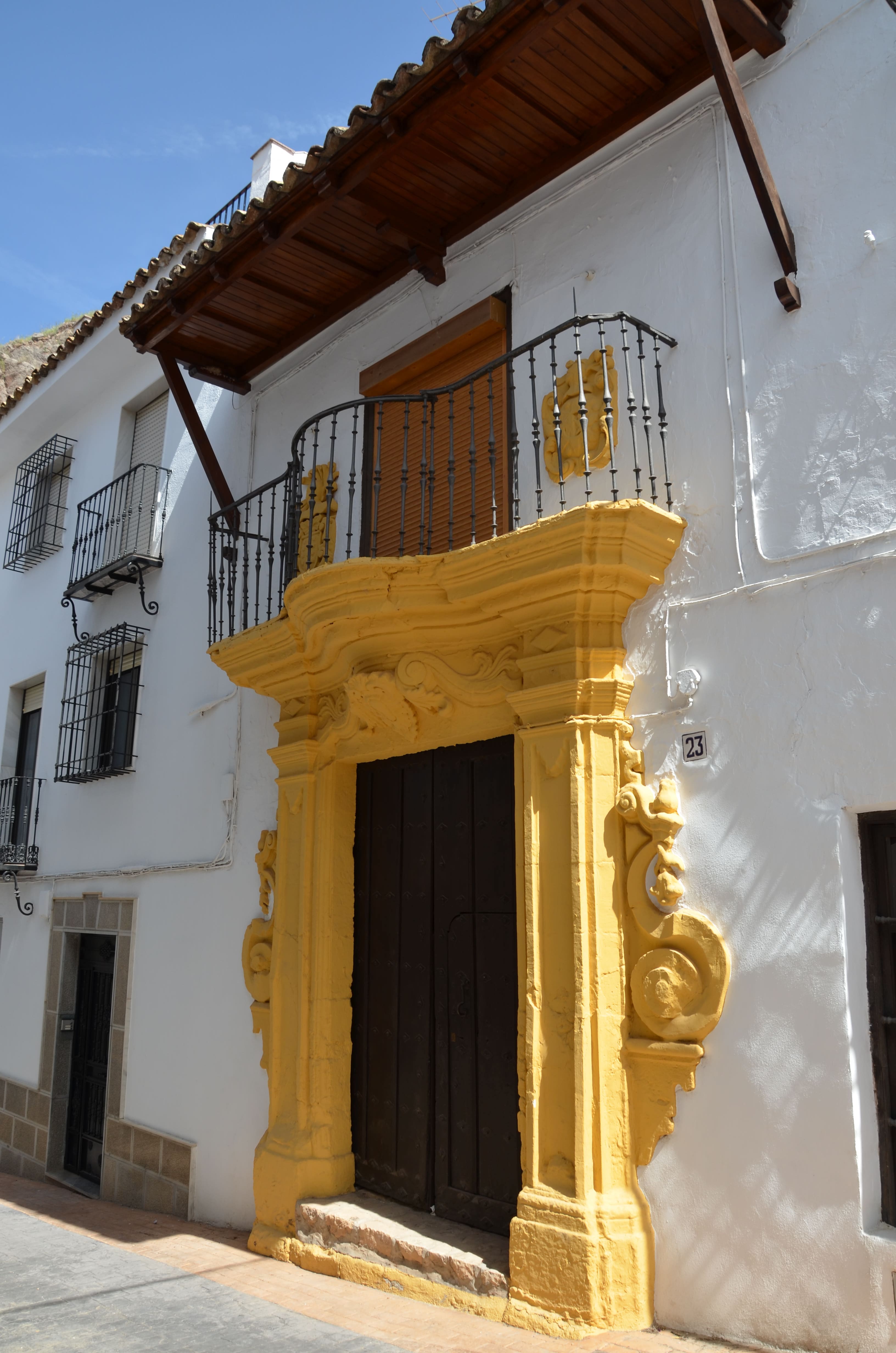 Detalle de una portada barroca pintada en amarillo, con molduras curvas y decoración tallada; sobre la puerta hay un balcón con barandilla de hierro negro.