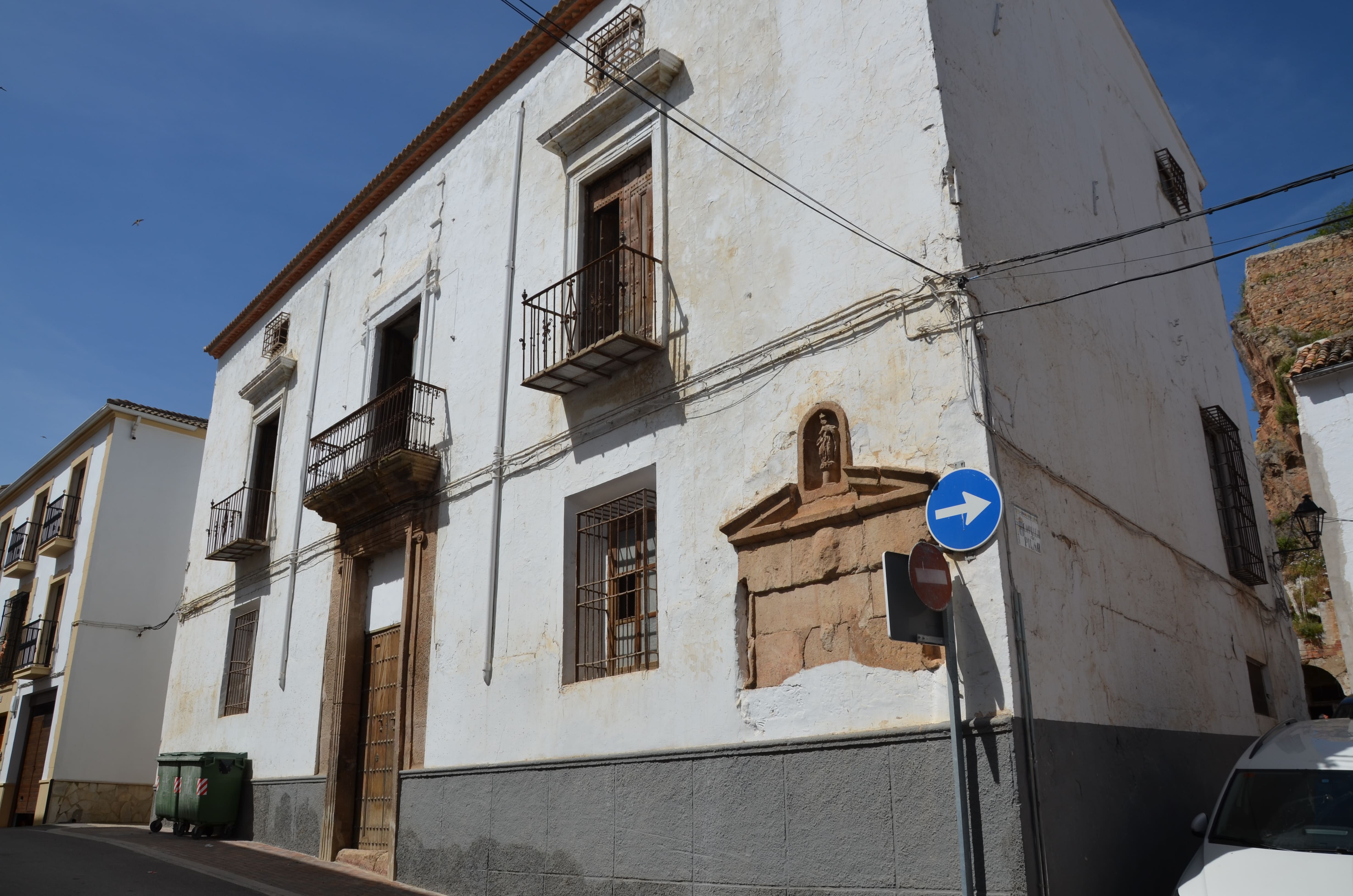 Fachada de una casa tradicional encalada en Cambil, vista en esquina desde la calle. El edificio es alto y rectangular, con varias ventanas protegidas con rejas y dos balcones de hierro forjado. Destaca una gran puerta de madera enmarcada por piedra, y en el lateral se aprecia un pequeño nicho con una hornacina que alberga una figura