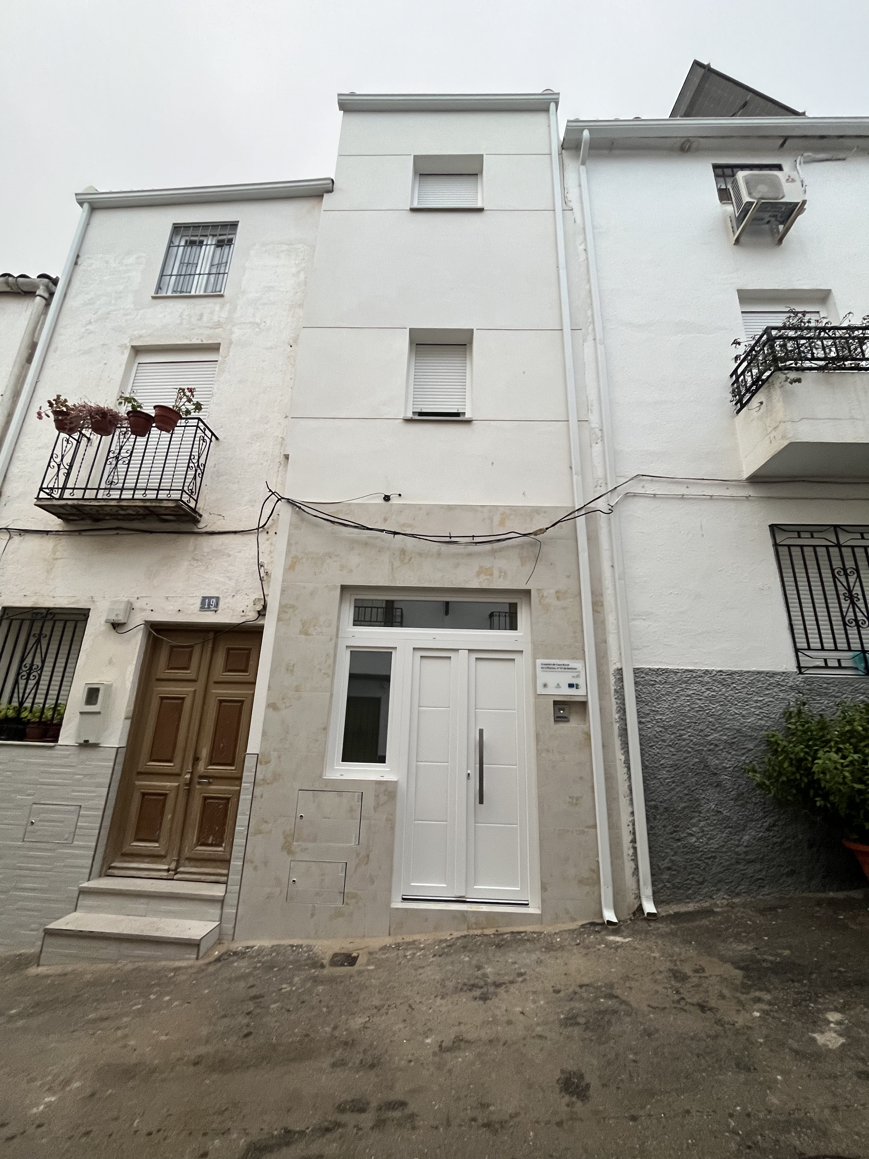 Fachada de una vivienda tradicional de tres plantas, con paredes blancas y detalles en piedra en la entrada. Se integra en una calle de pendiente, acompañada de balcones y elementos típicos del caserío andaluz.