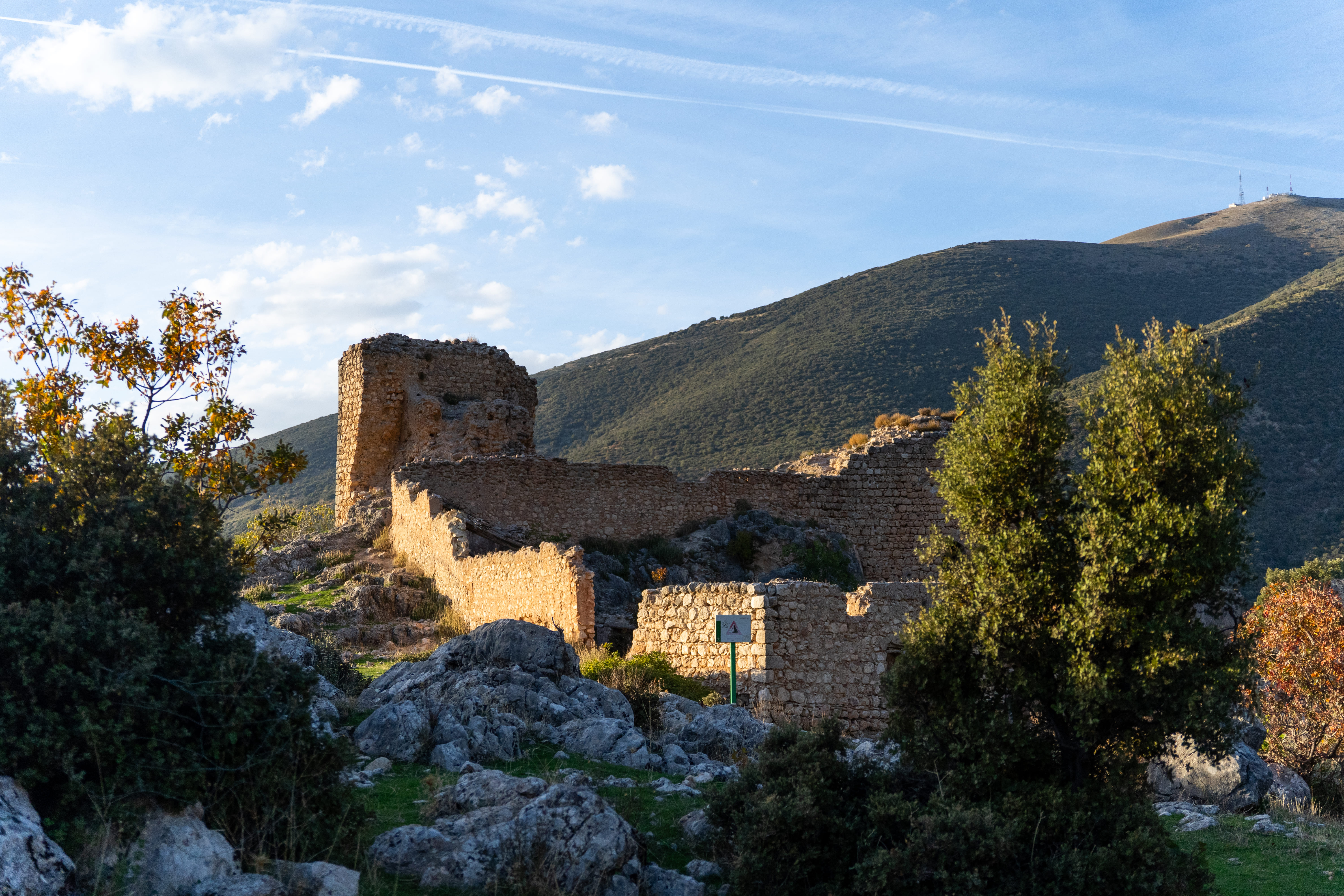 Ruinas de una antigua construcción de piedra situadas entre rocas y vegetación, en la ladera de una montaña. La luz cálida resalta los muros y el relieve del entorno, bajo un cielo azul con algunas nubes dispersas.