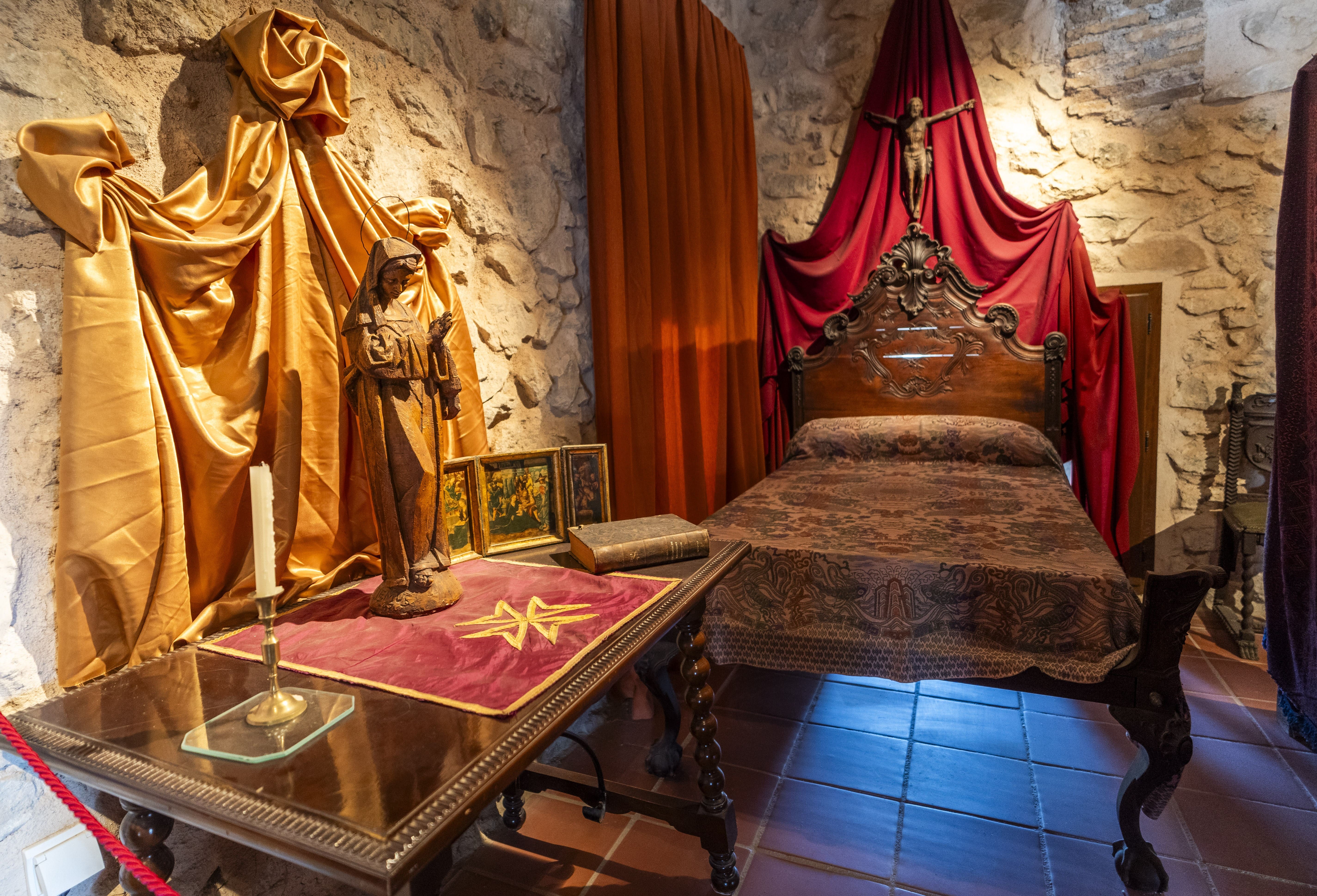 Recreación interior de una estancia histórica del castillo, con cama de madera, tejidos decorativos, una figura religiosa y mobiliario ambientado en época antigua.