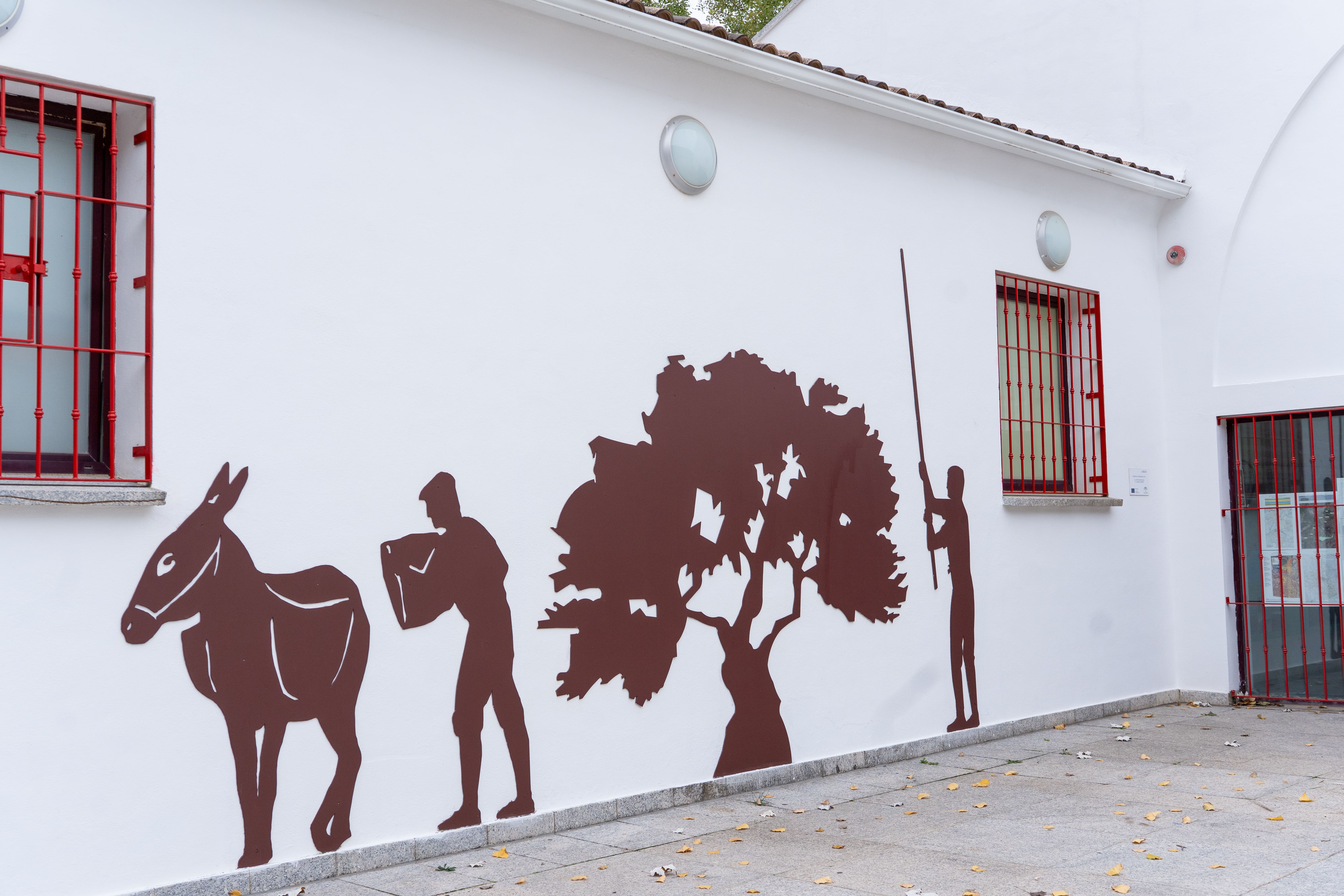 Silueta metálica en una pared blanca que representa escenas tradicionales del campo (un burro, una persona y un árbol), con ventanas enrejadas