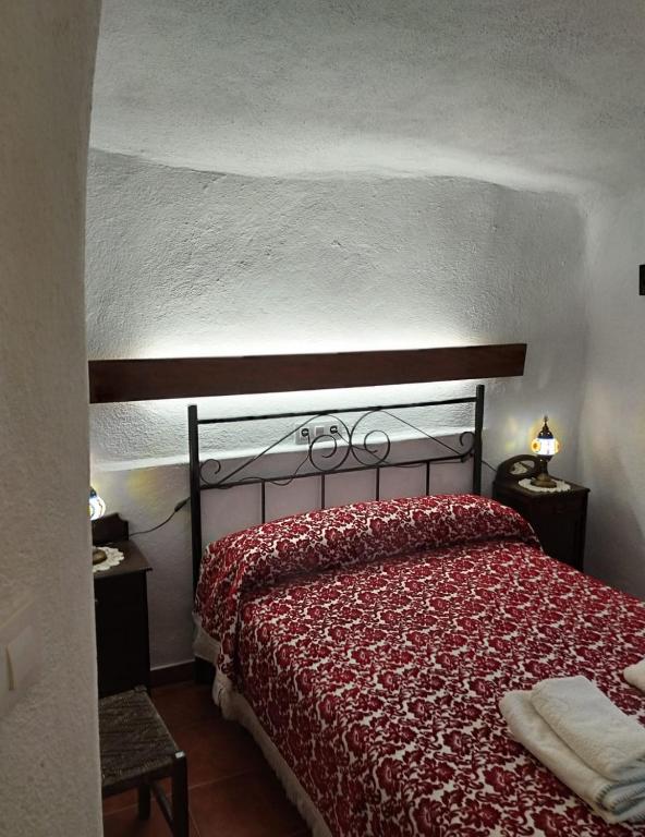 Dormitorio rústico y acogedor con paredes encaladas de formas orgánicas, que evocan una casa cueva. La cama, con colcha floral roja y cabecero de forja, se acompaña de luz cálida y detalles sencillos que invitan al descanso.