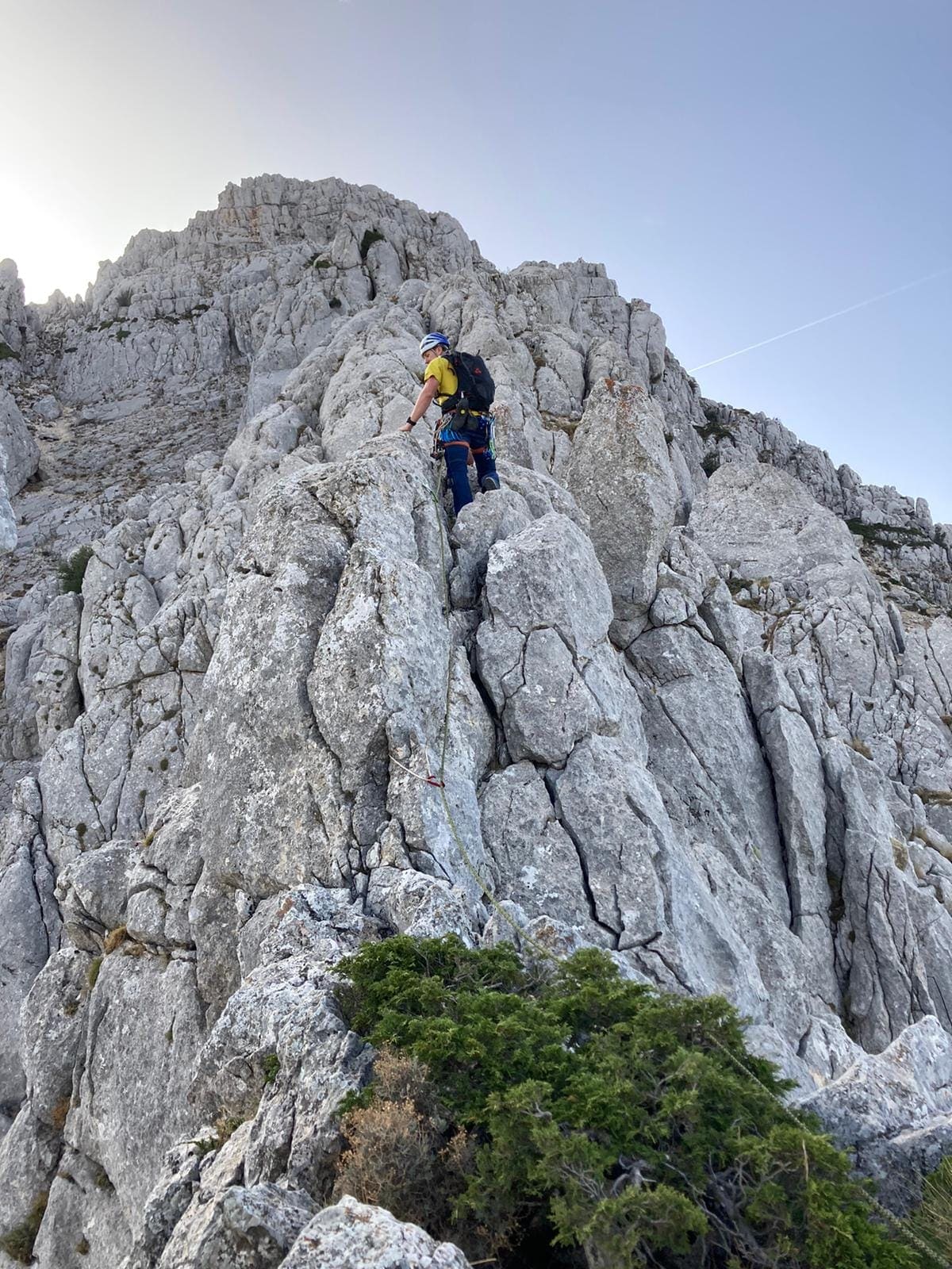 Persona con casco, mochila y material de escalada ascendiendo por una cresta rocosa de color claro en alta montaña. El terreno es abrupto y pedregoso, con una cima rocosa al fondo.