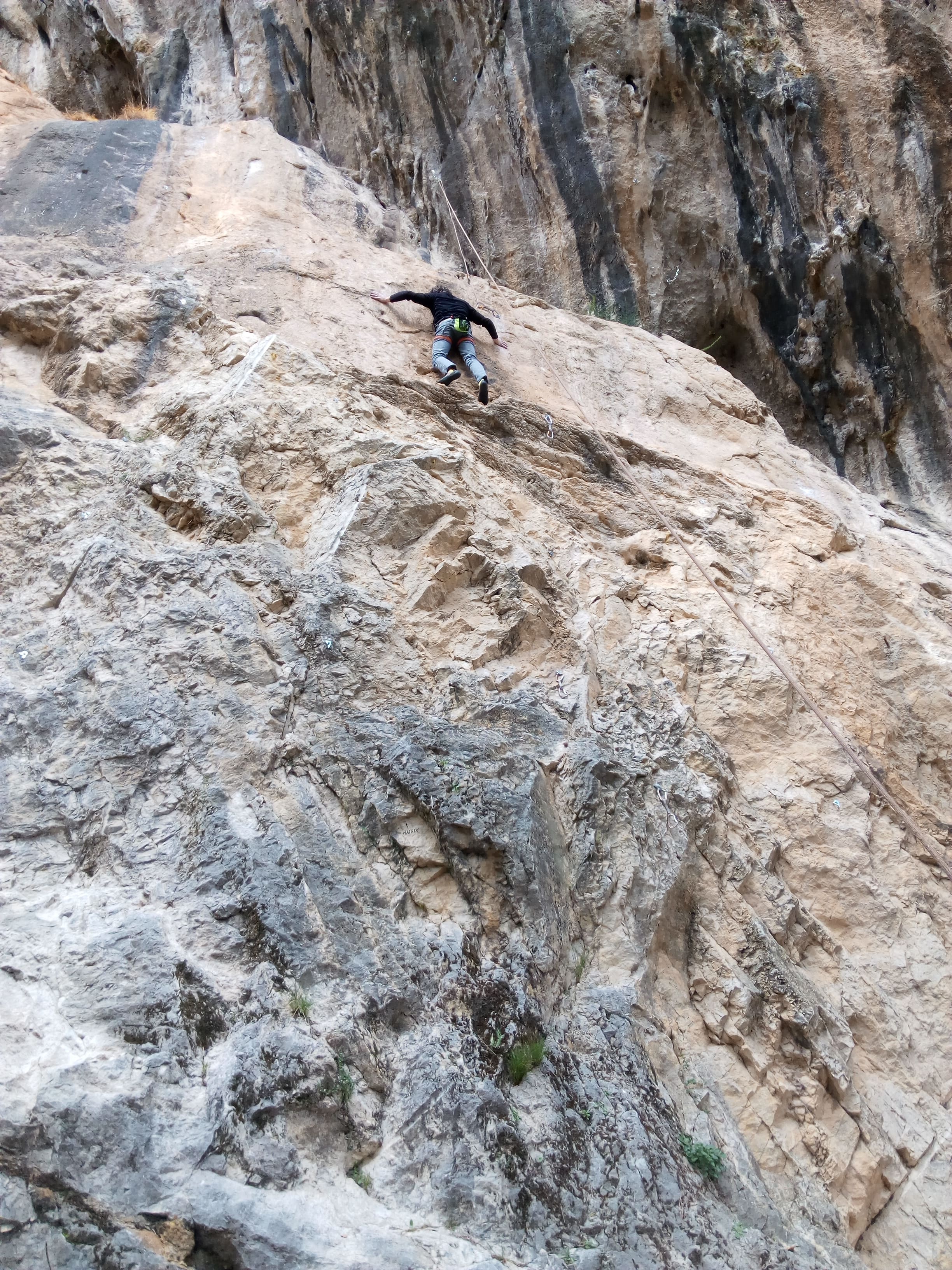 Persona escalando una gran pared de roca inclinada, sujeta con cuerda de seguridad. La superficie rocosa muestra distintos tonos beige y gris, con textura irregular y aspecto desafiante.