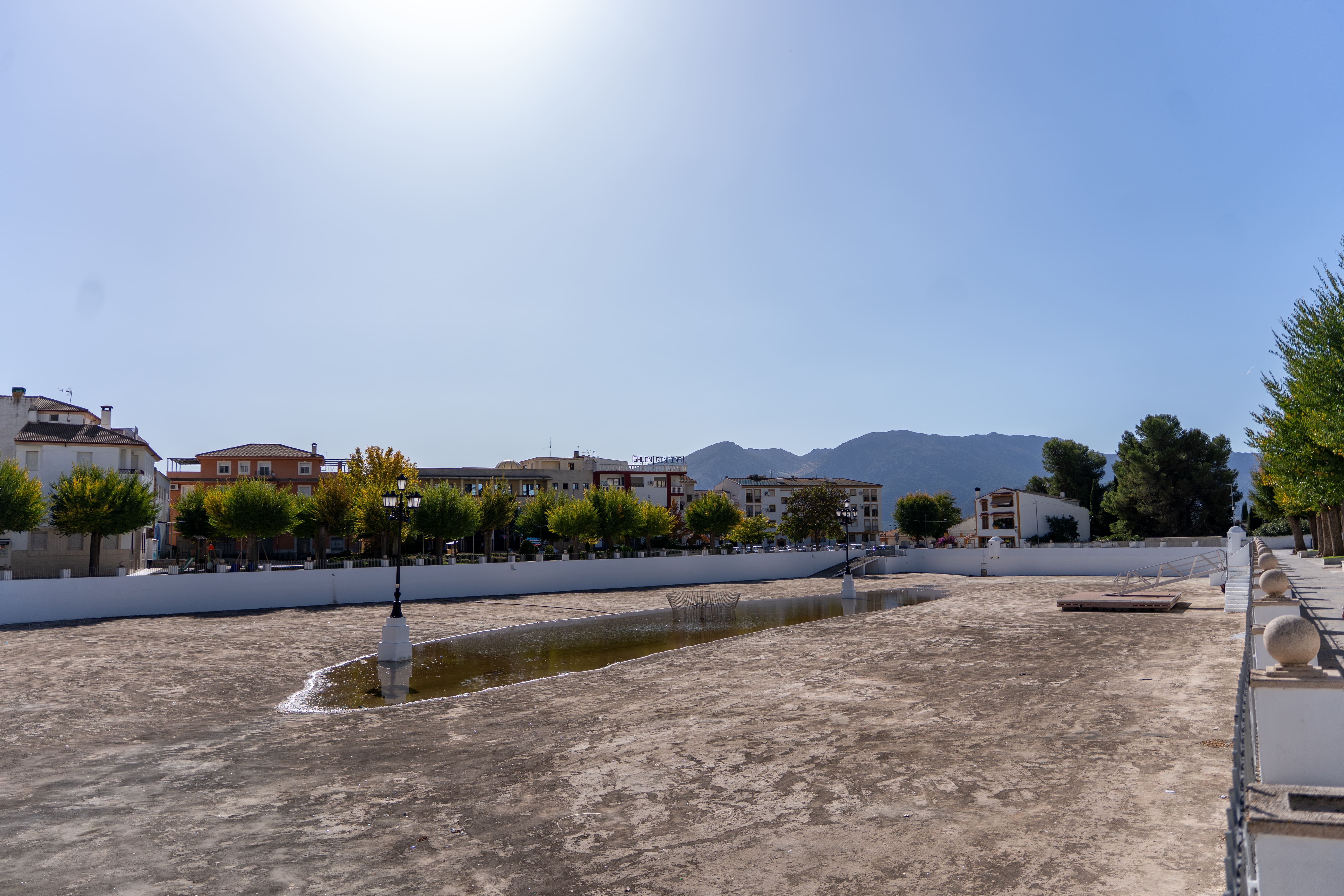 Espacio urbano abierto con una gran lámina de agua muy reducida, rodeado de un paseo, arbolado y edificios. Al fondo se recortan las montañas bajo un cielo despejado, en una escena amplia y luminosa.
