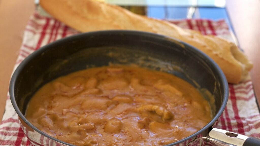 Gachas tradicionales servidas en una sartén, con una textura espesa y cremosa de color anaranjado. Al fondo aparece una barra de pan sobre un paño de cuadros rojos y blancos, acompañando este plato de cuchara típico de la cocina tradicional.