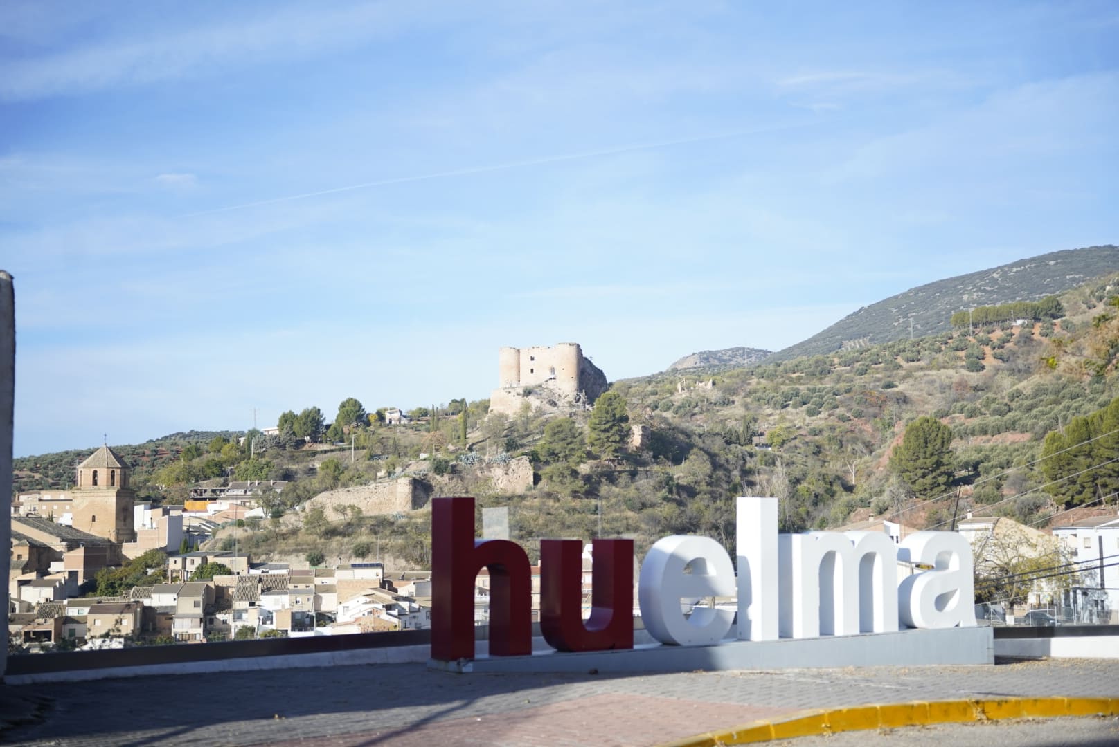 Vista panorámica de Huelma, con el castillo de los Duques de Alburquerque dominando la colina al fondo y la iglesia del municipio visible entre las casas del casco urbano. En primer plano aparece el cartel con el nombre del pueblo, desde un mirador que ofrece amplias vistas del paisaje de olivares y montañas de Sierra Mágina.