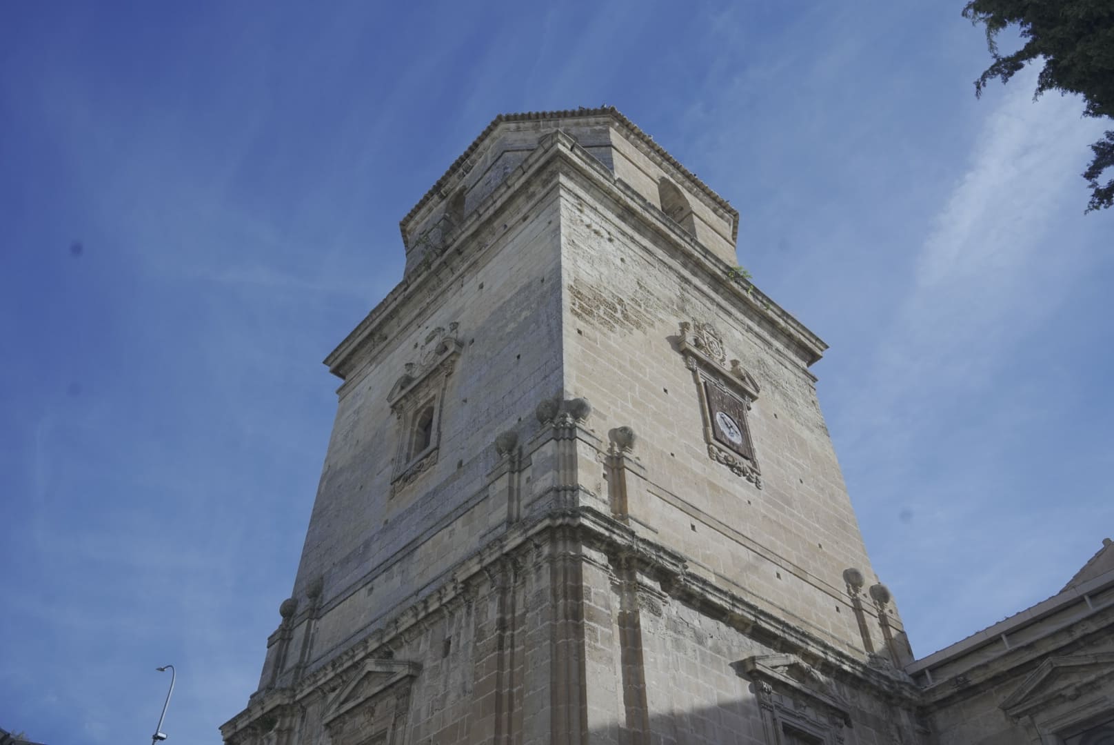 Vista en contrapicado de la torre campanario de la Iglesia de la Inmaculada Concepción, construida en piedra y organizada en varios cuerpos. La luz resalta los detalles arquitectónicos renacentistas y el reloj situado en uno de los niveles de la torre.