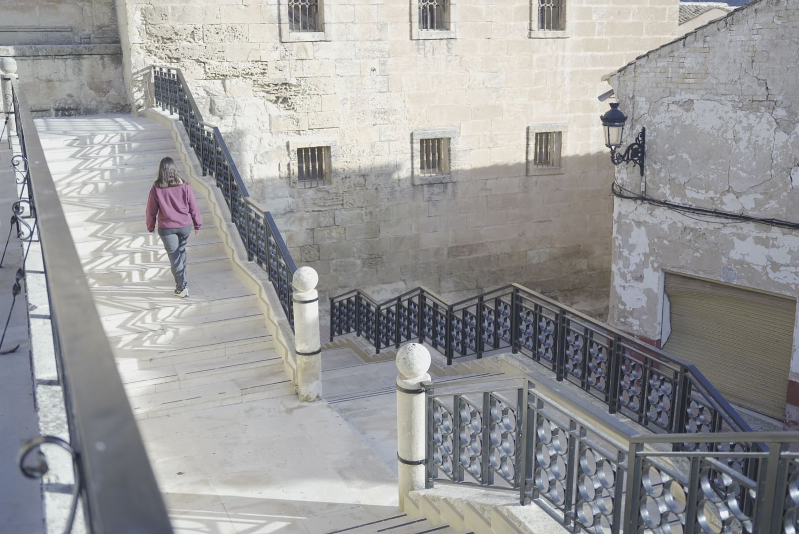 Acceso lateral mediante una escalinata con barandillas de hierro que conecta distintos niveles del entorno de la iglesia. Una persona desciende por el paseo elevado junto a los muros de piedra del templo, integrados en el entramado urbano del casco histórico.
