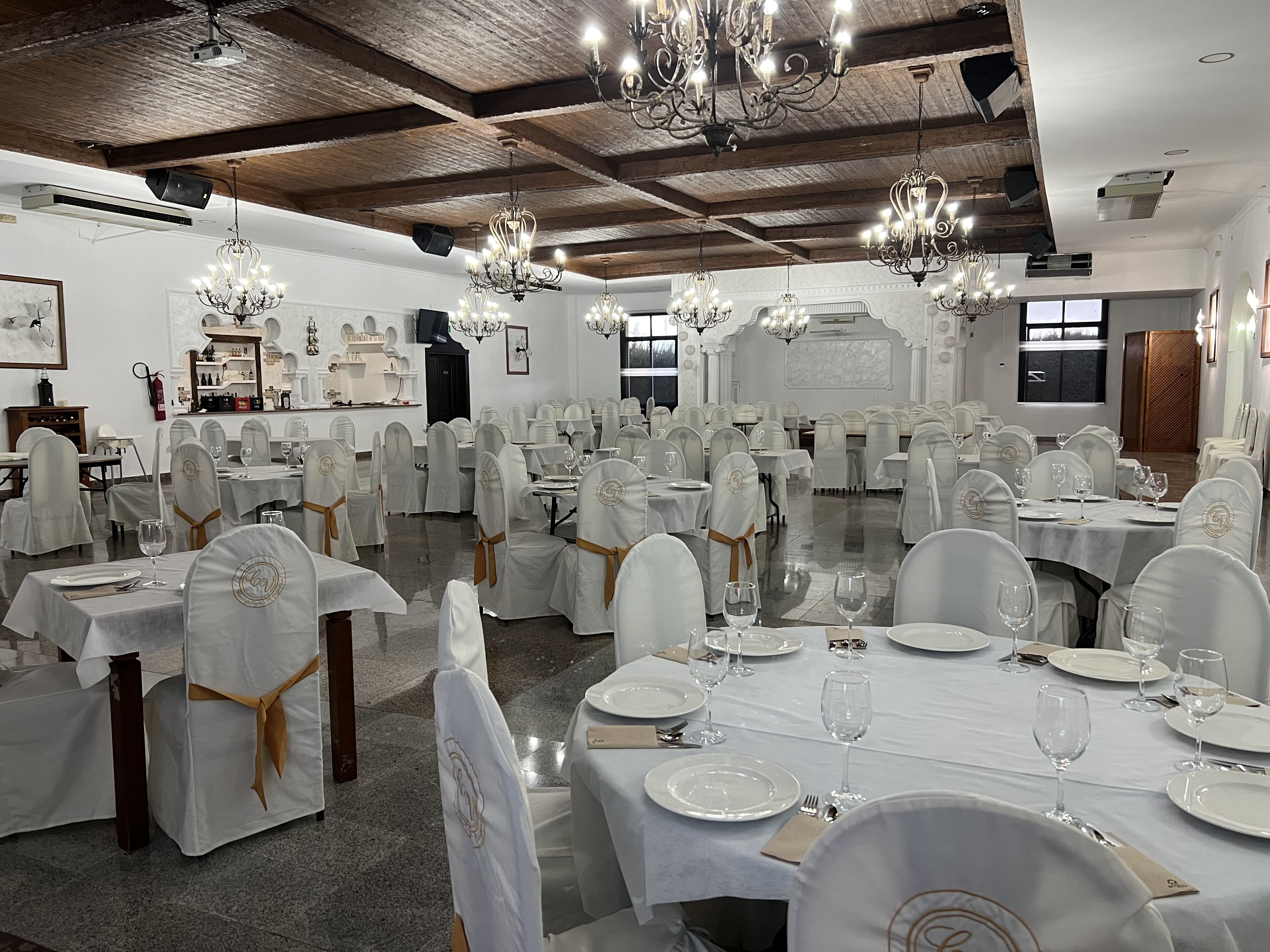 Amplio salón-comedor preparado para eventos o celebraciones, con mesas vestidas de blanco, sillas con fundas y lámparas de araña colgando del techo de madera.