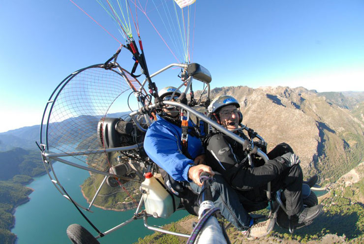 Dos personas sobrevuelan un paisaje de montaña y embalse a bordo de un paramotor.