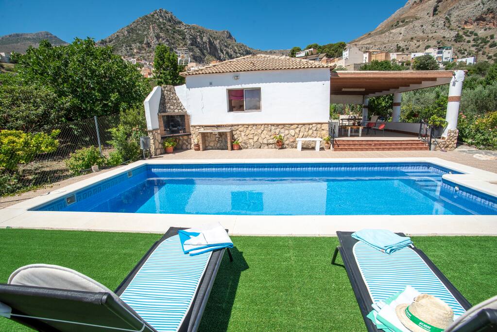 Piscina privada en primer plano con tumbonas, junto a una casa rural de fachada blanca y detalles en piedra. Al fondo, un porche cubierto y un entorno de montaña que enmarca la escena, transmitiendo tranquilidad y disfrute al aire libre.