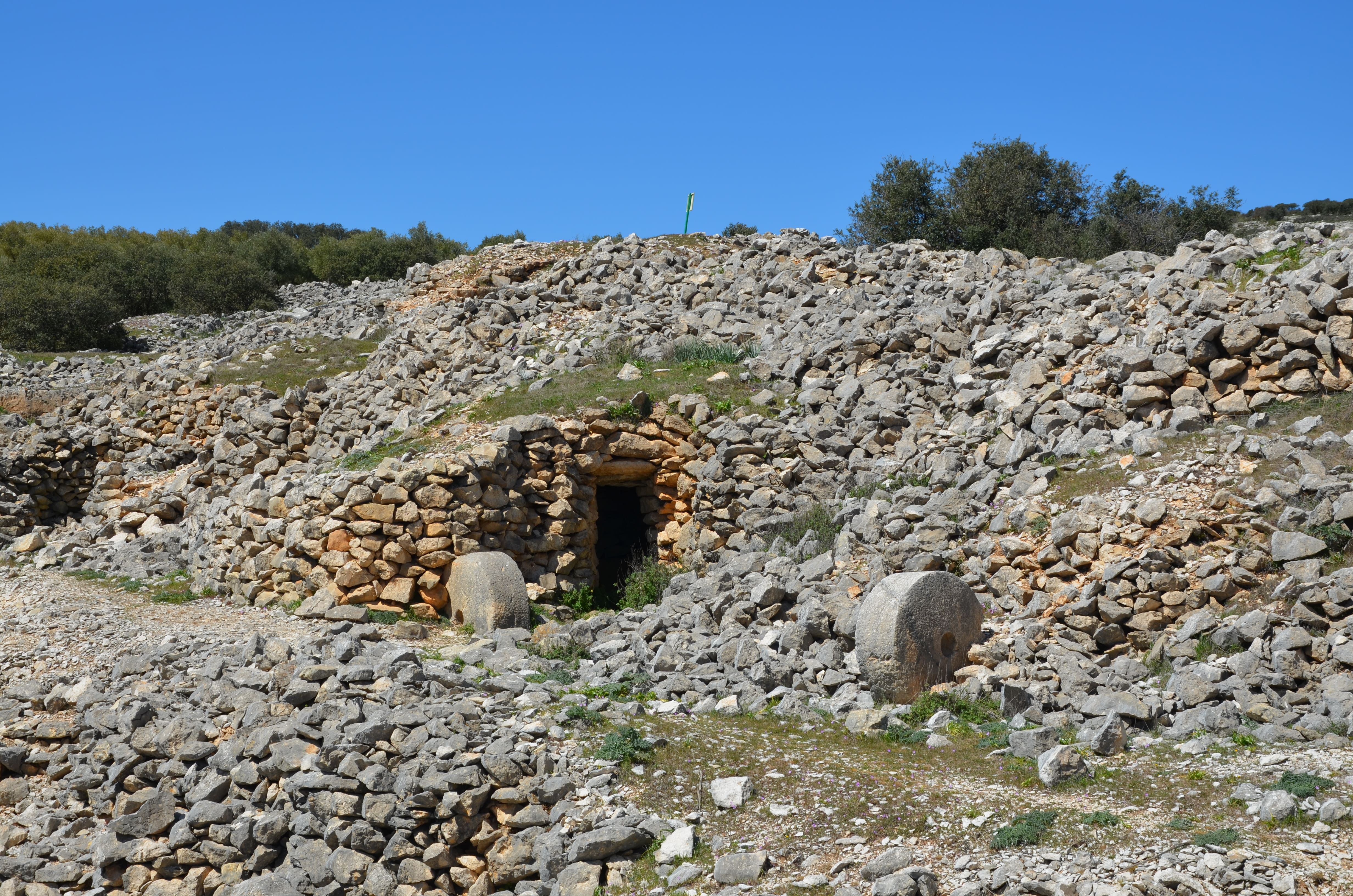 Vista de un paisaje rocoso en el paraje de Los Caracoles, en Sierra Mágina. Se aprecian antiguas construcciones de piedra seca integradas en la ladera, con una entrada oscura y varias piedras de molino junto a la estructura.