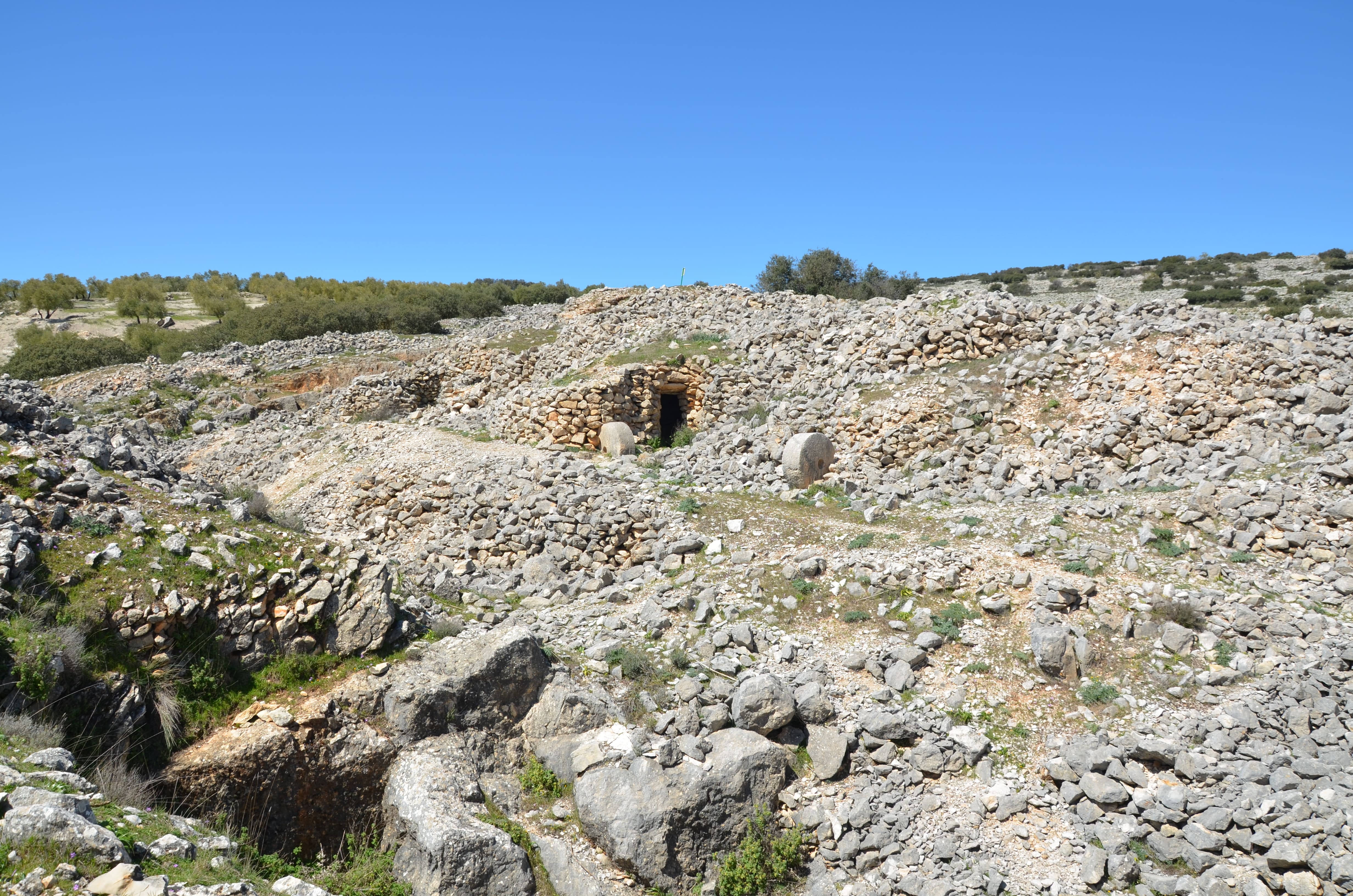 Paisaje de piedra seca en Los Caracoles, con muros y refugios tradicionales construidos con piedras del terreno. La imagen muestra el conjunto integrado en la montaña, rodeado de vegetación y bajo un cielo despejado.