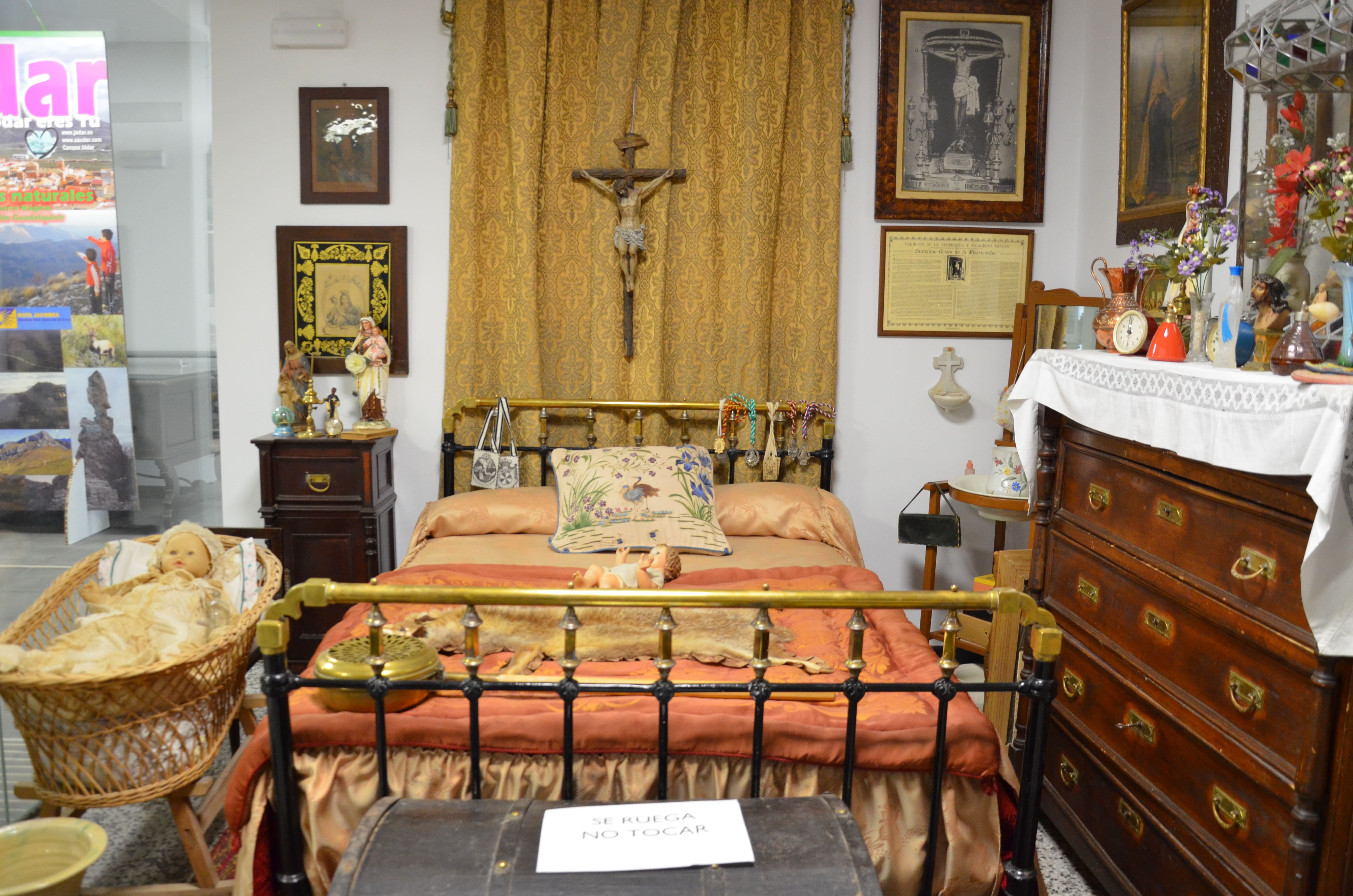 Recreación de una habitación antigua con cama de hierro, cuna de mimbre, cómoda de madera, objetos religiosos, cuadros y elementos decorativos tradicionales. El espacio muestra cómo era una estancia doméstica del pasado.
