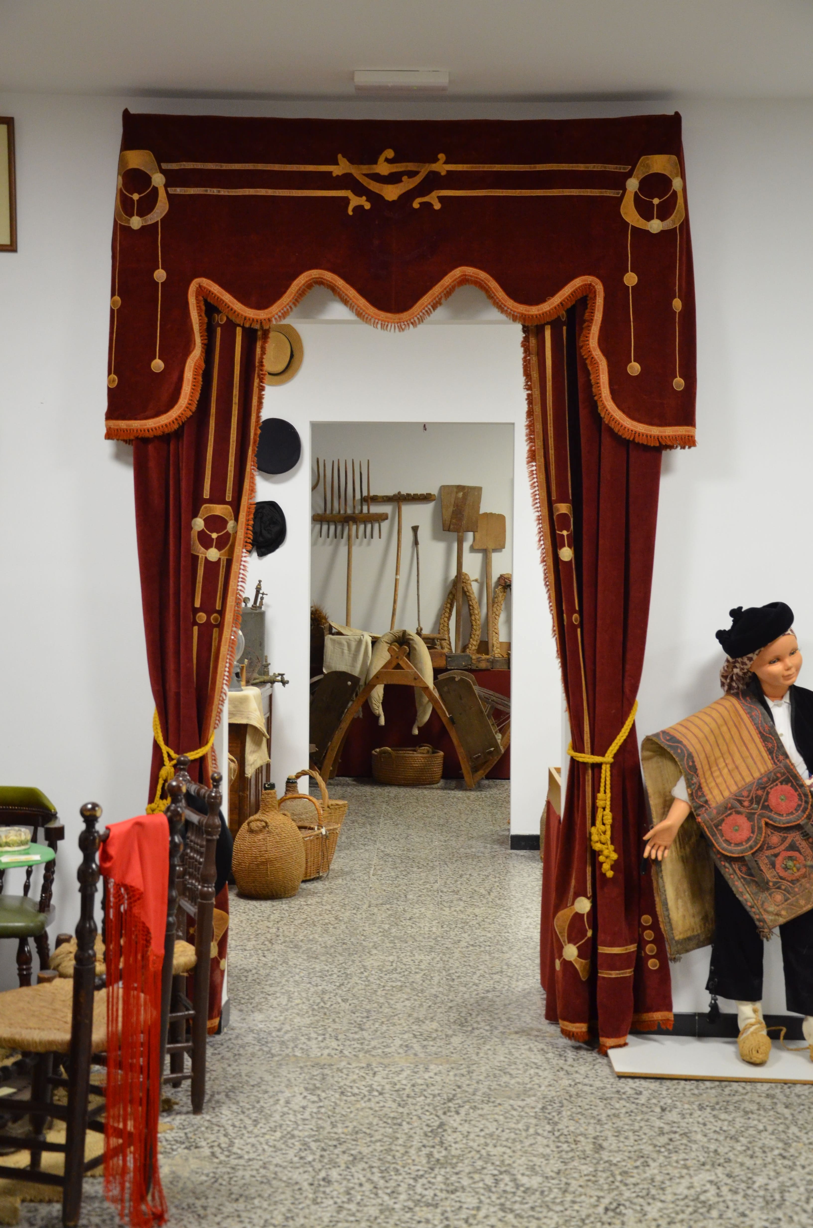 Sala expositiva con una recreación de ambiente tradicional. Se observan cortinas rojas, mobiliario antiguo, objetos de uso cotidiano, herramientas agrícolas y una figura vestida con indumentaria popular.