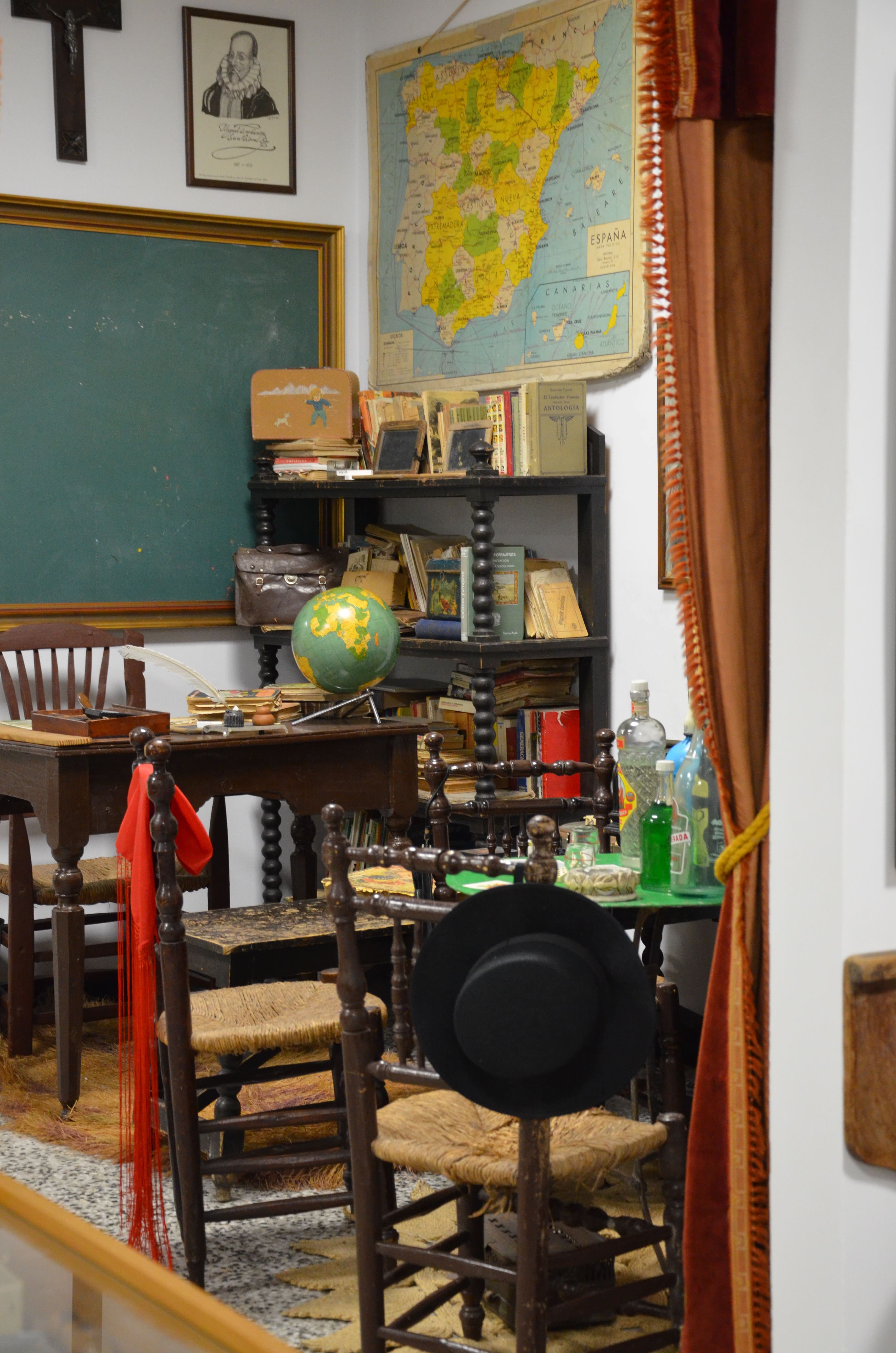 Vista de una sala museográfica con mobiliario escolar y objetos antiguos. Aparecen pupitres, sillas, libros, una pizarra, un globo terráqueo y un mapa de España, recreando un aula de época.