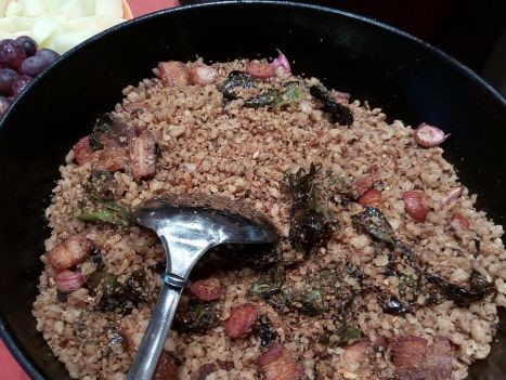Migas tradicionales servidas en una sartén negra, acompañadas con trozos de panceta, chorizo y pimientos fritos. En el centro aparece una cuchara metálica, y al fondo se aprecia fruta como acompañamiento, típico contraste de este plato popular.
