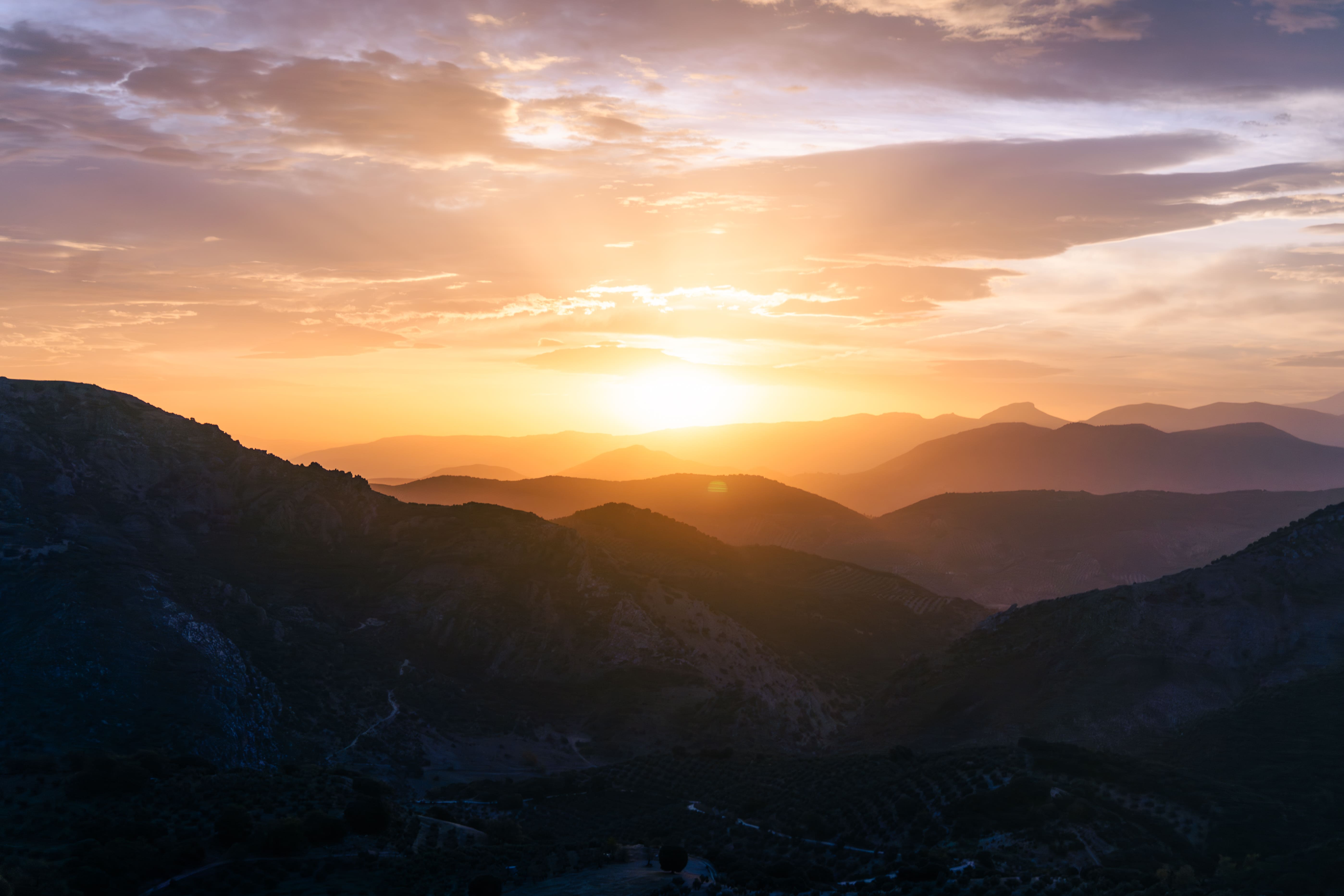 La fotografía muestra un atardecer sobre las sierras de Noalejo, donde el sol ilumina el horizonte con tonos anaranjados y dorados. Las montañas se superponen en silueta creando un paisaje natural de gran belleza.