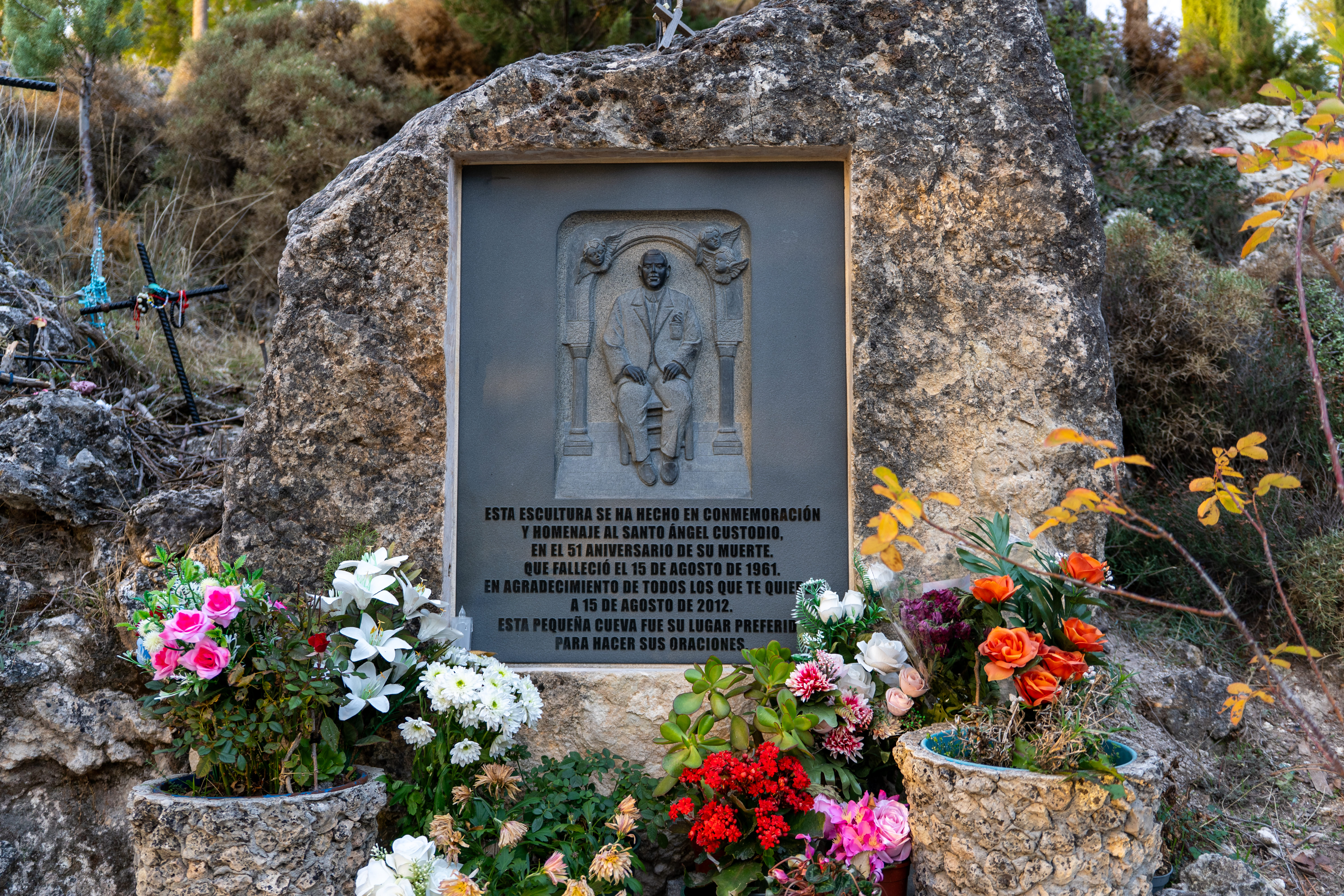 La imagen presenta un monumento dedicado al Santo Ángel Custodio, tallado en una piedra natural e integrado en el entorno. A su alrededor hay flores y elementos de devoción que reflejan el carácter espiritual del lugar.