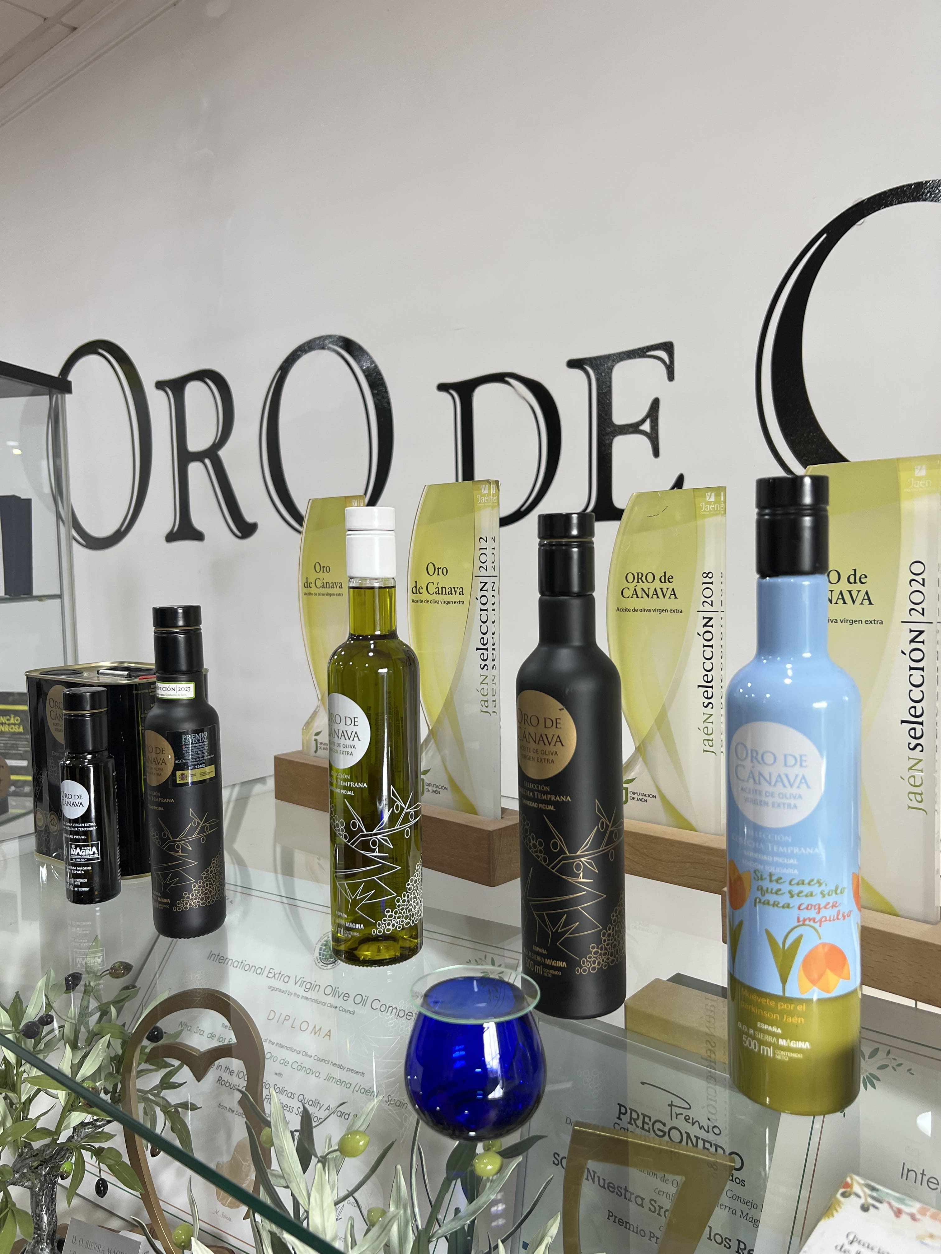 Detalle de una vitrina con diferentes envases de aceite de oliva virgen extra Oro de Cánava, junto a diplomas y elementos decorativos vinculados al olivar.