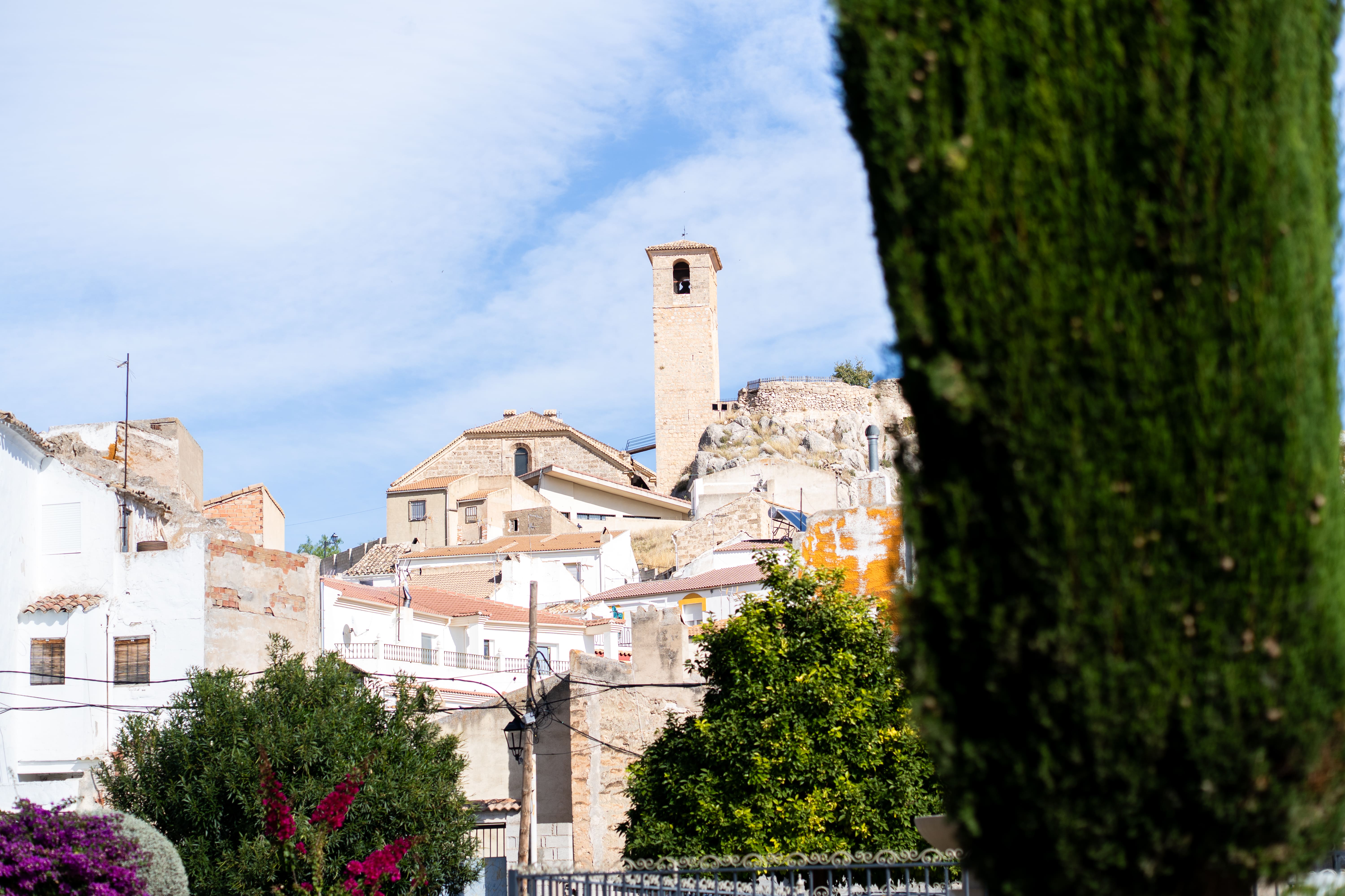 Vista parcial de un núcleo urbano de casas blancas y tejados de teja, con una torre campanario de piedra que sobresale entre los edificios. La vegetación en primer plano enmarca la escena y aporta profundidad al paisaje urbano.