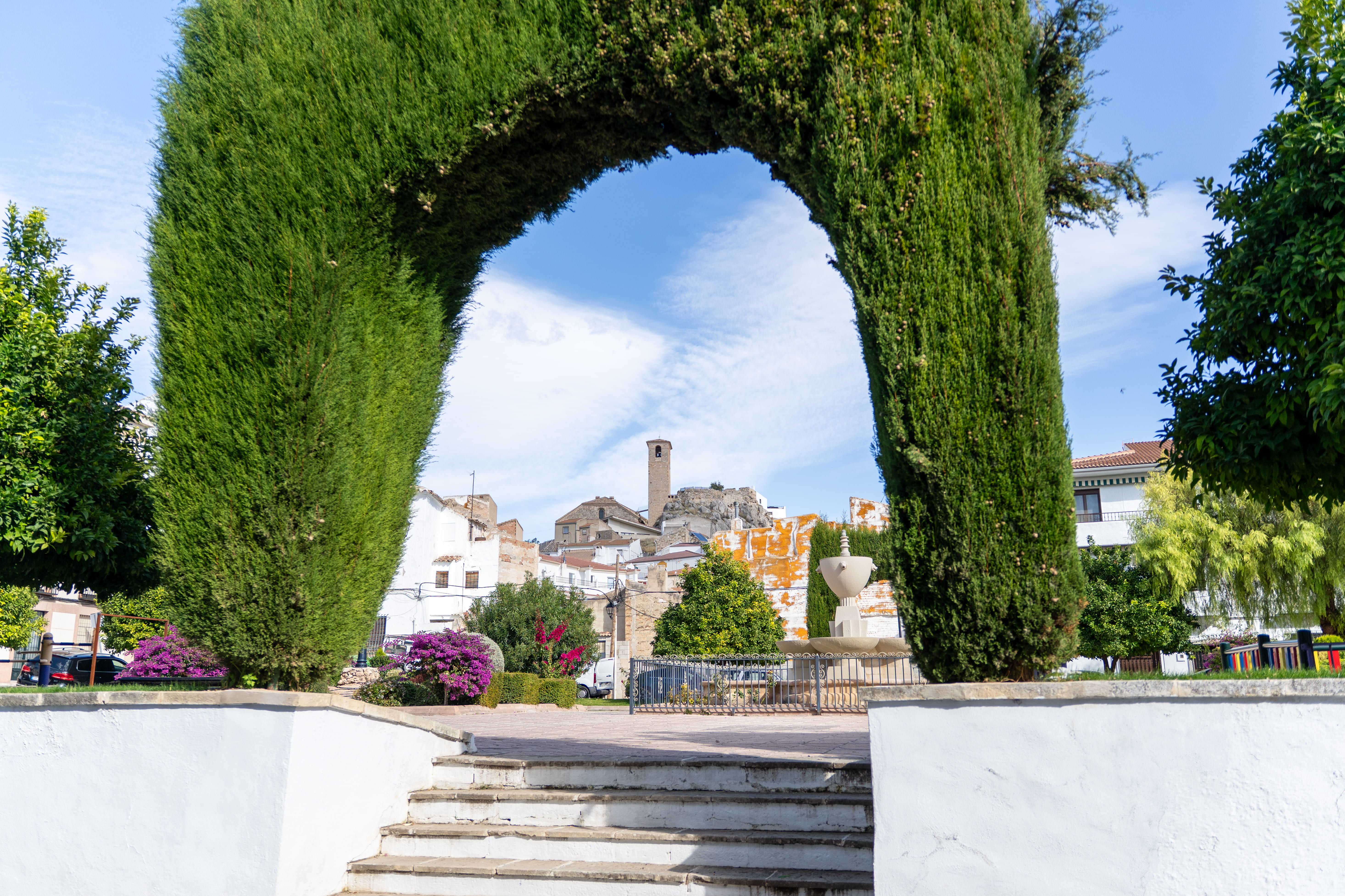 Arco vegetal formado por altos cipreses recortados que enmarca una plaza con escalinata, jardines y una fuente. Al fondo se distinguen casas blancas y una torre de piedra que sobresale sobre el conjunto urbano.