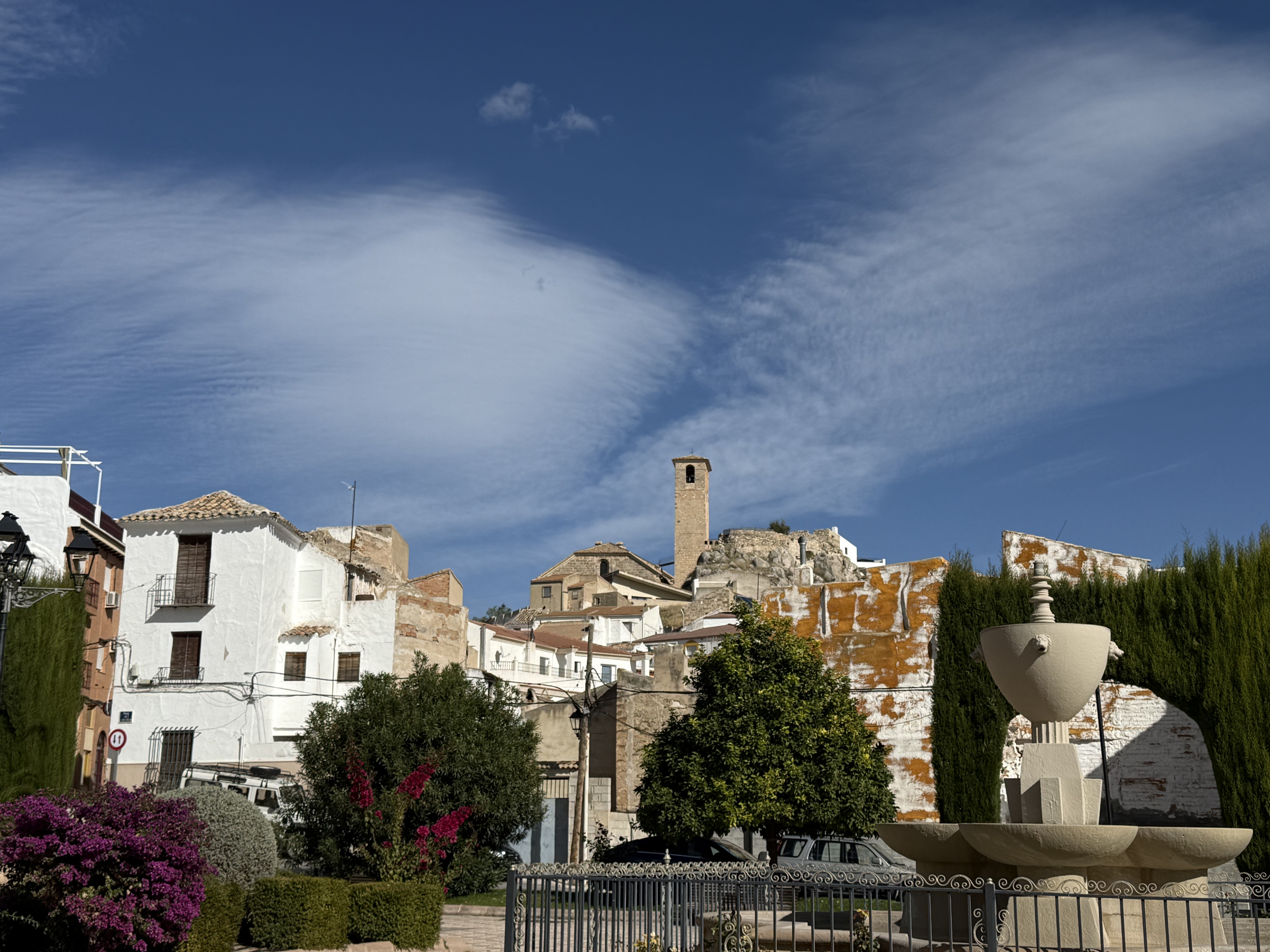Un pintoresco pueblo de casas blancas y tejados tradicionales se extiende bajo un cielo azul intenso, decorado por nubes suaves y alargadas. En el centro destaca una antigua torre sobre una colina, mientras una fuente y jardines aportan encanto y tranquilidad a la escena.